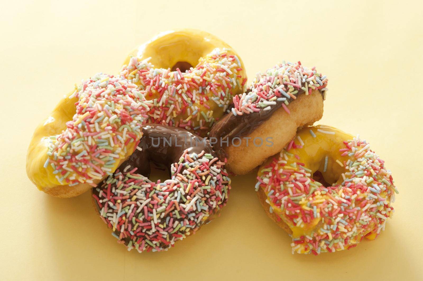 Pile of assorted fresh ring donuts or doughnuts glazed with chocolate, lemon or orange icing decorated with colorful sprinkles on a pale yellow background