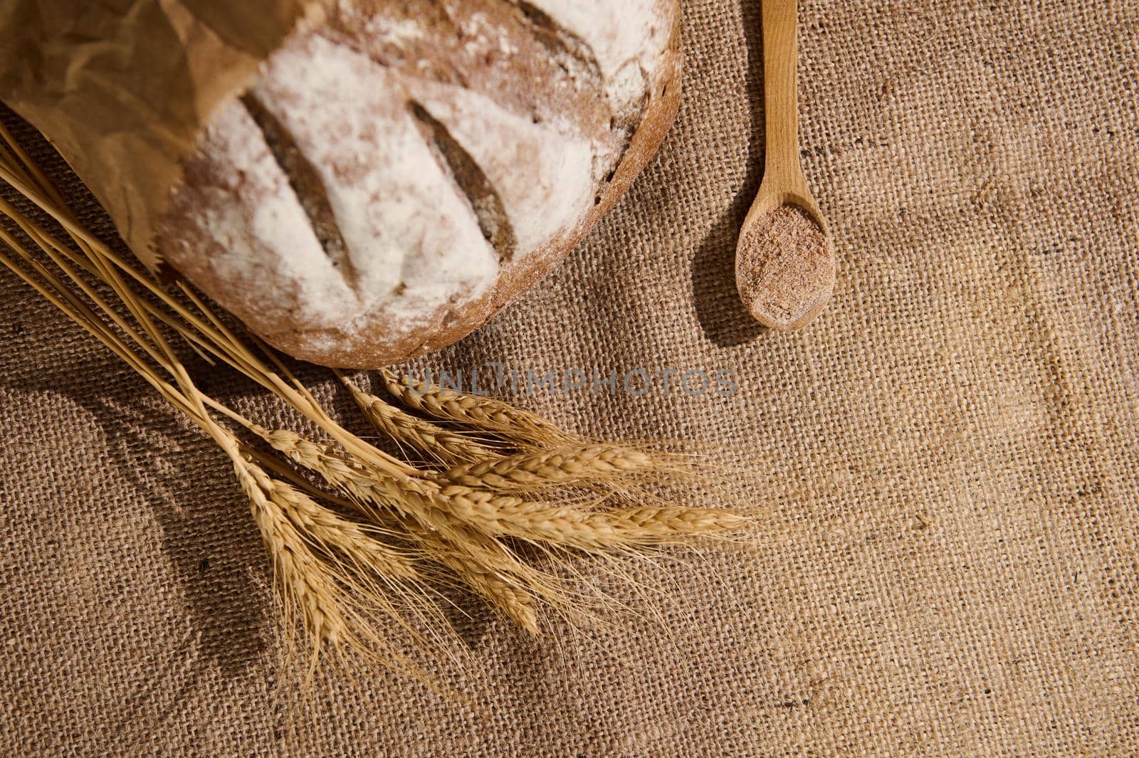 A loaf of whole grain bread sprinkled with flour, spikelets of wheat and a wooden spoon with bran on sacklock tablecloth by artgf