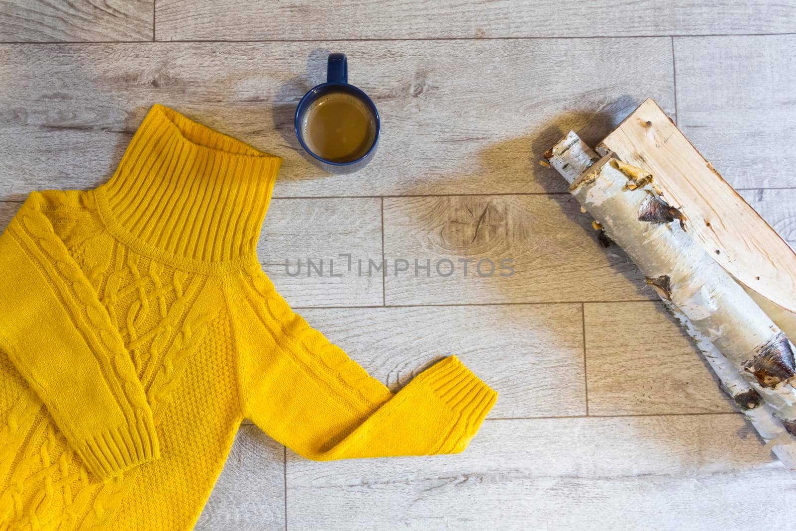 A yellow sweater on a gray, wooden background next to a blue mug of hot drink. and birch firewood..