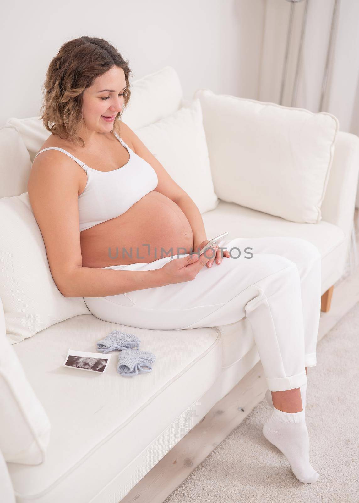 Pregnant woman sits on a white sofa and holds a smartphone. Fetal ultrasonic screening. by mrwed54