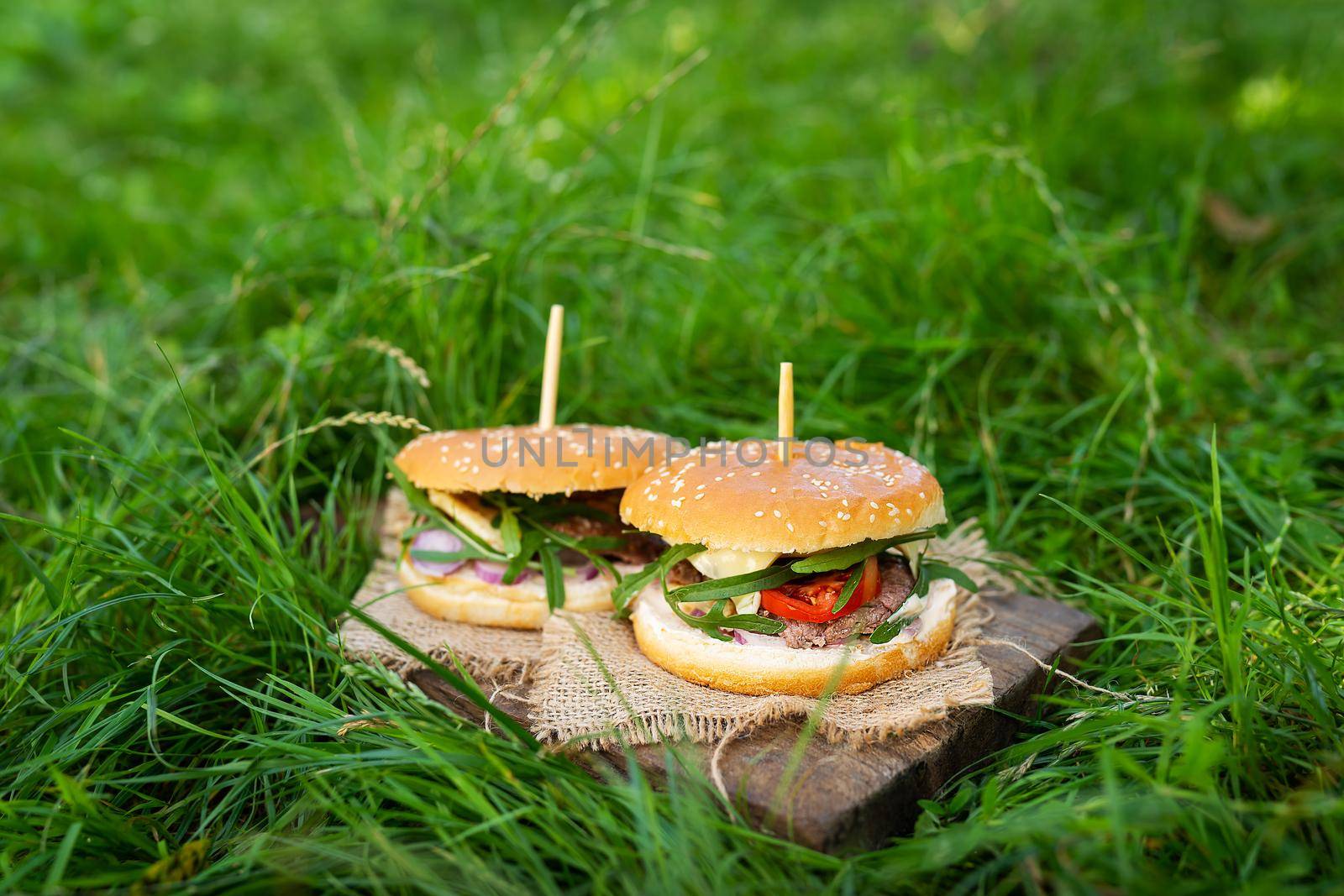 A juicy and tasty burger stands on a board on the green grass in the street outdoors. Close-up, sunny day