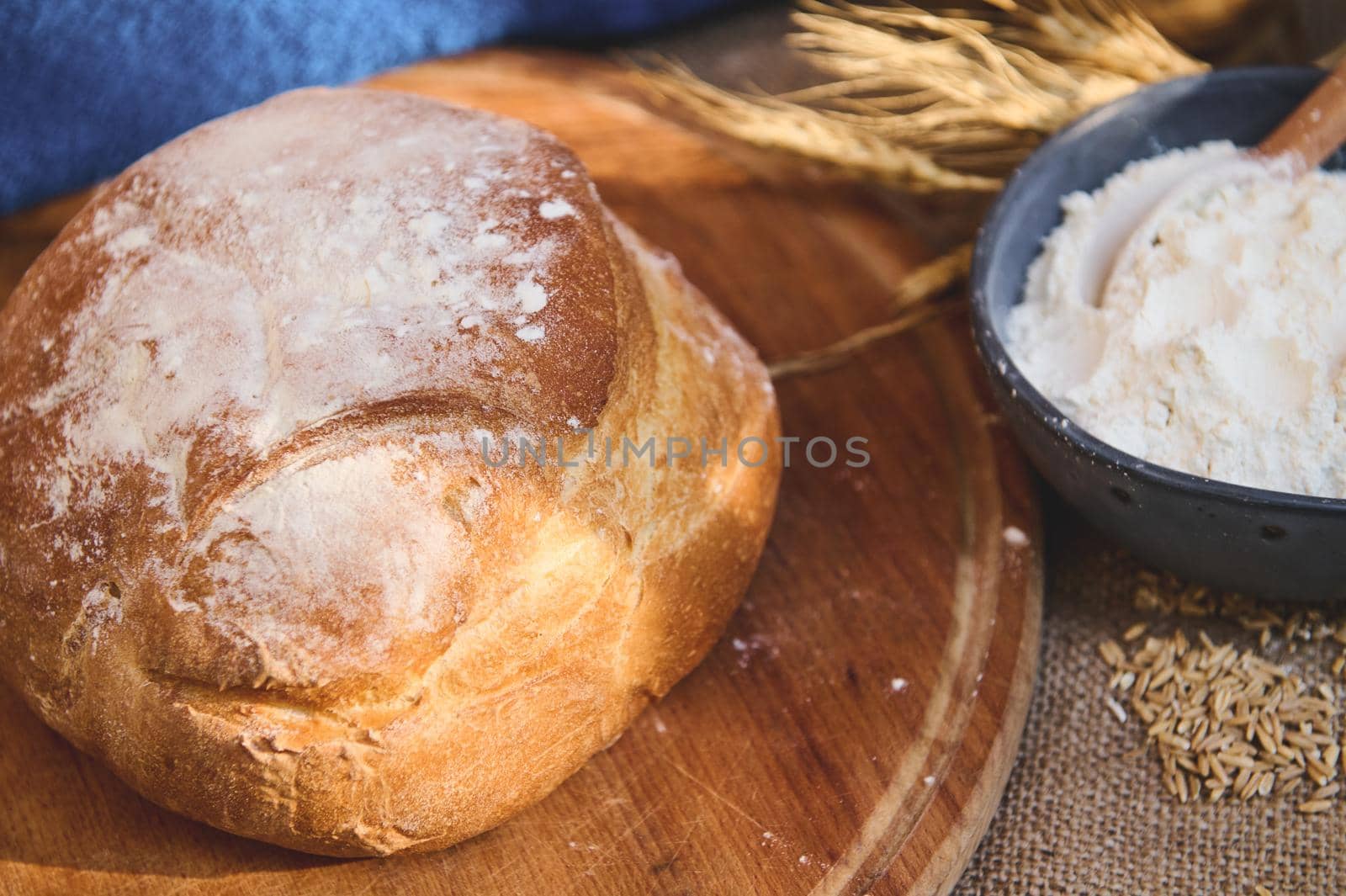 Selective focus. Overhead view of a loaf of homemade whole grain sourdough bread on a wooden board, against blurred blue ceramic bowl with white flour and spikelets of wheat. Artisanal bakery store