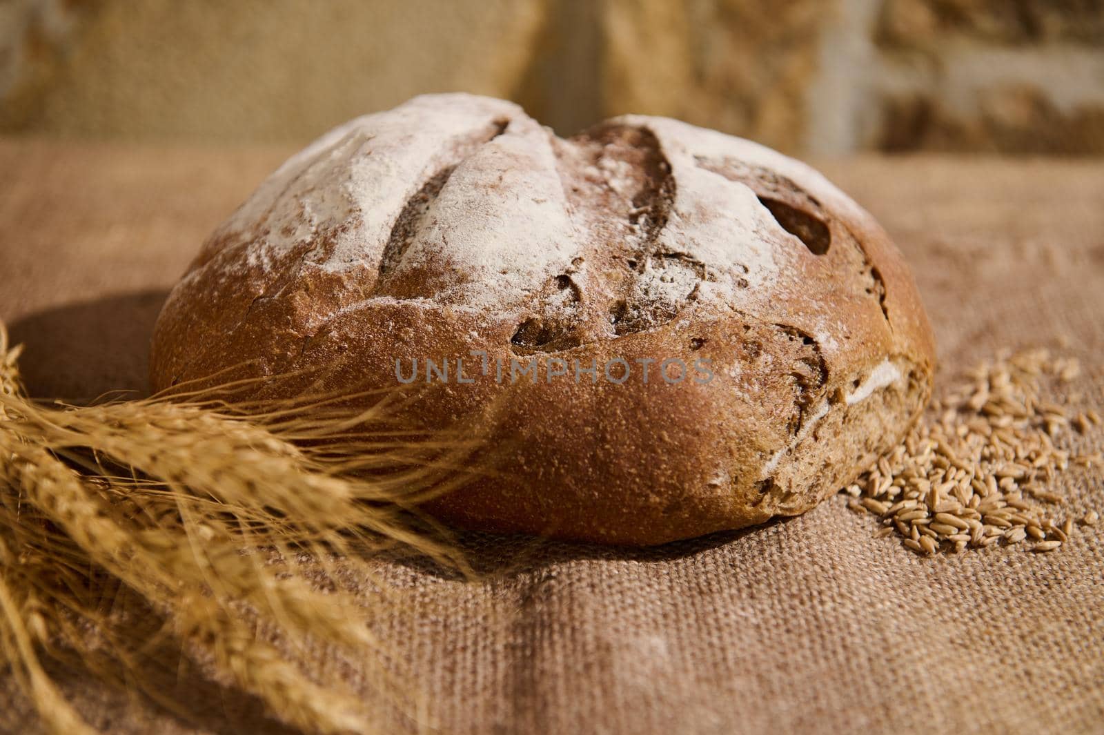 Close-up of a homemade wholesome multigrain sourdough bread, grains scattered on the burlap surface, and blurred wheat ears on the foreground. Artisanal bakery. Healthy baking items enriched in fiber