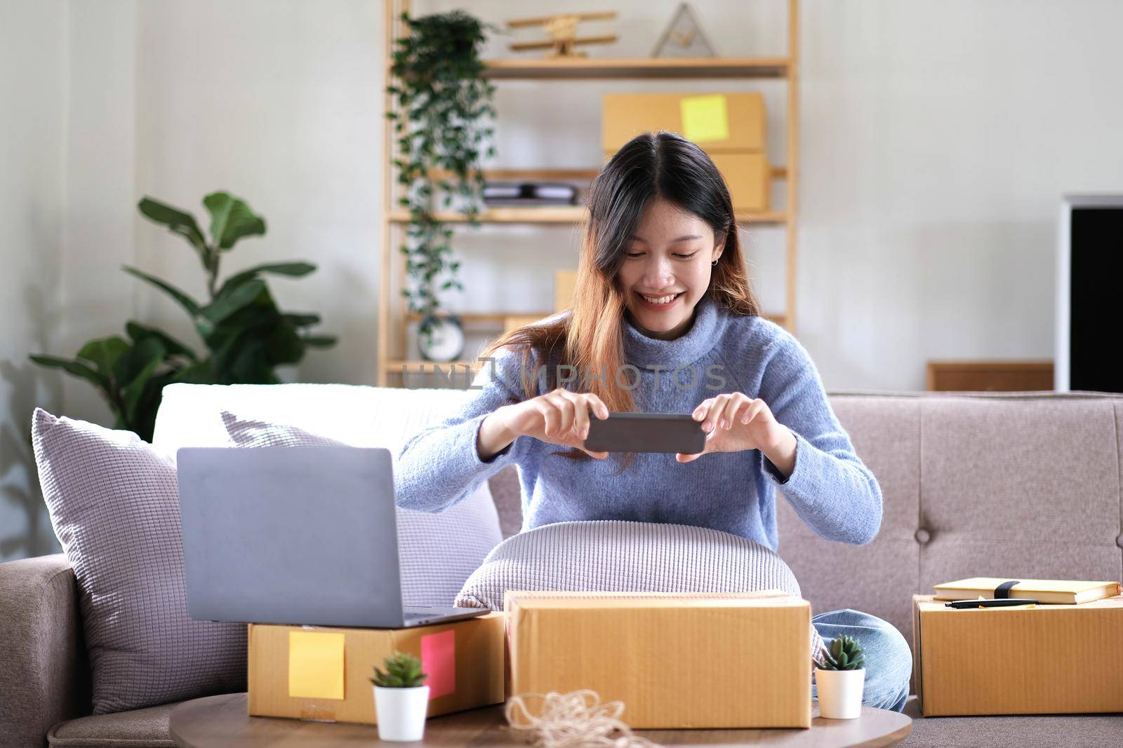 Asian woman photographing parcel boxes with smartphone to deliver goods to customers online on the Internet, starting a small business owner's e-commerce concept