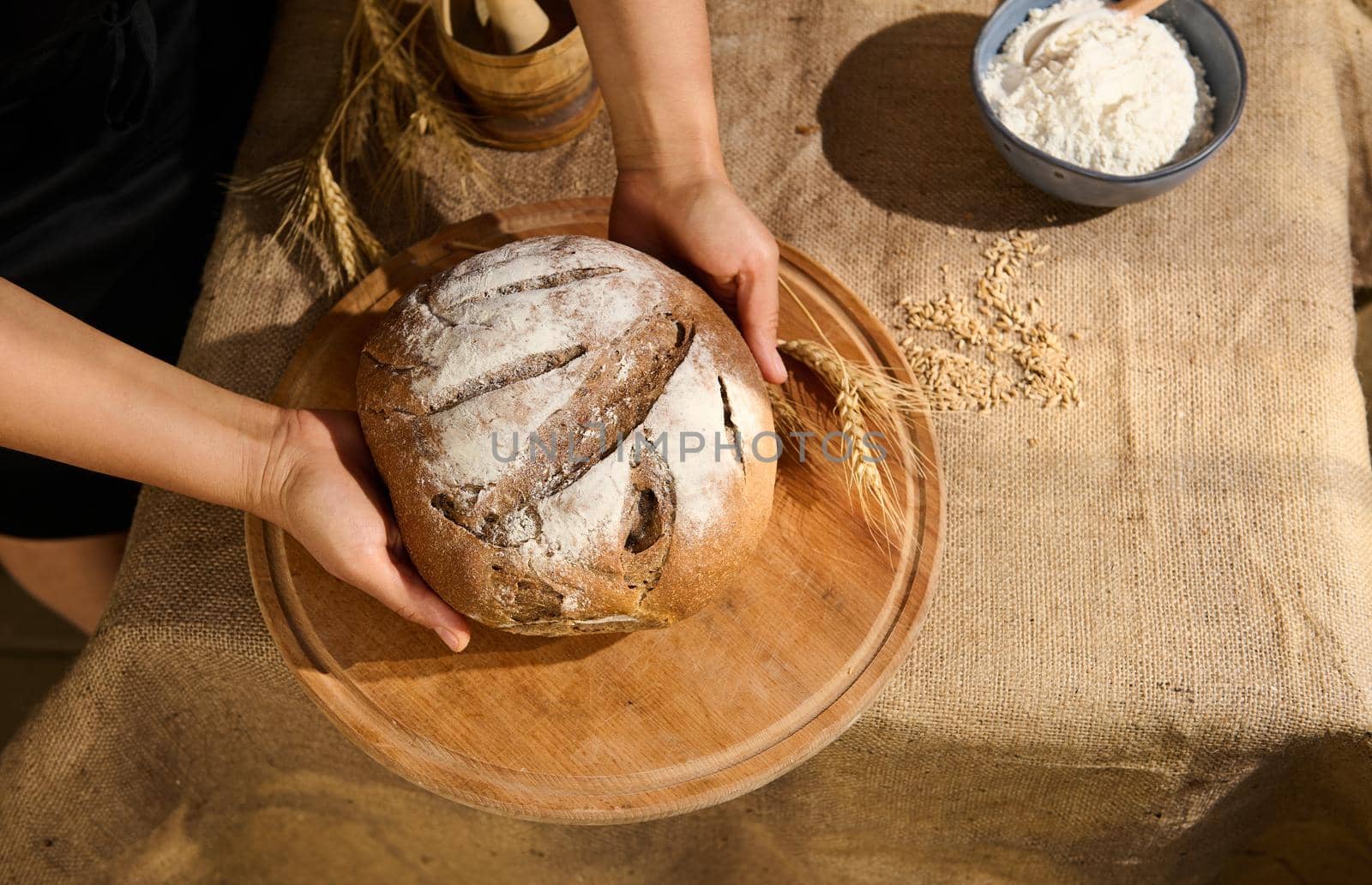 View from above. Baker confectioner holds a loaf of warm traditional homemade whole grain sourdough wheat bread, enriched in dietary fiber, puts it on a wooden board, in the artisanal family bakery