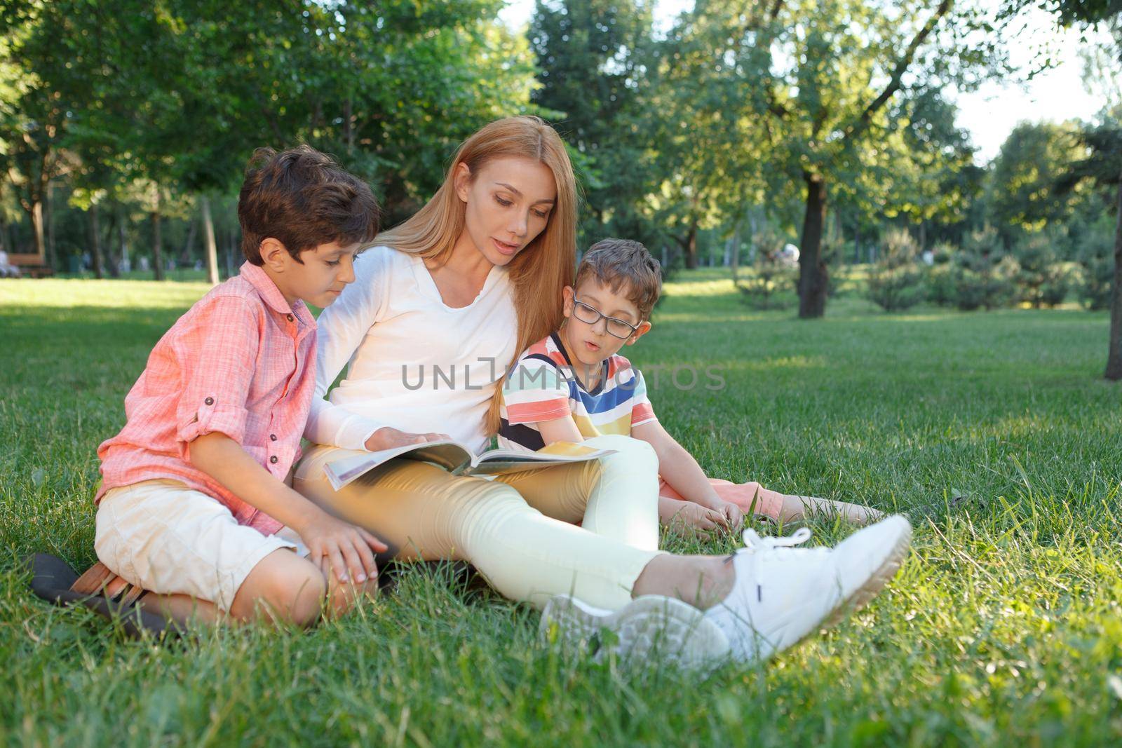 Two little schoolboys reading a book with their female teacher outdoors in the park
