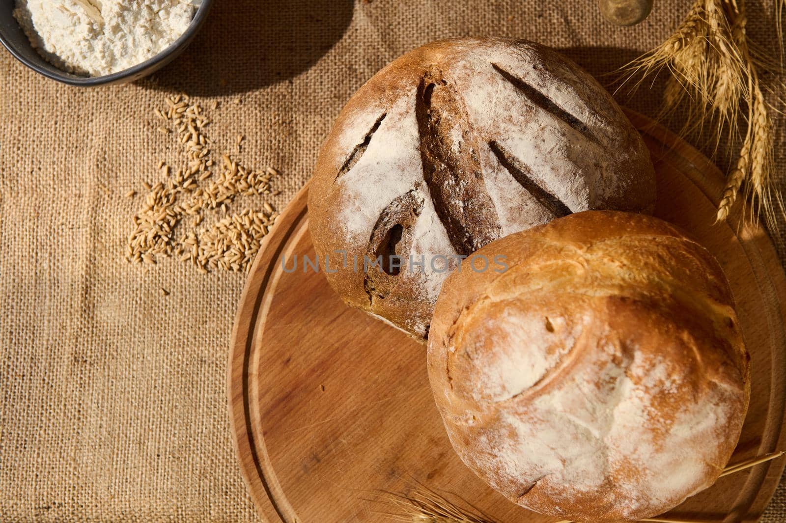 Top view. Loaves of fresh homemade bread on a wooden board with scattered grains and wheat spikelets on a burlap tablecloth. Still life with wholesome bakery, enriched in dietary fiber. Copy ad space