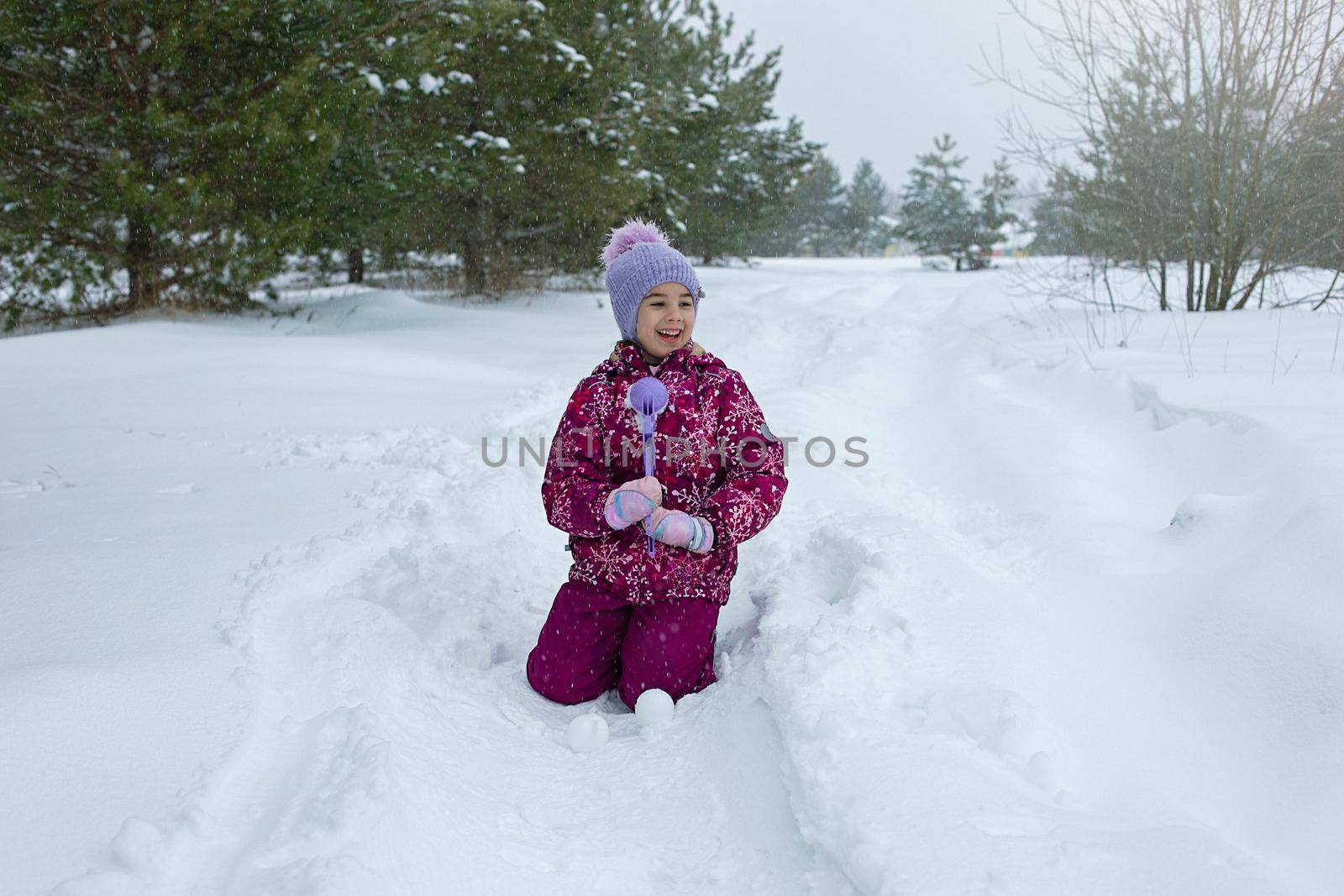 A little girl , sits in the snow, makes snowballs using a plastic sculpting tool. Close up