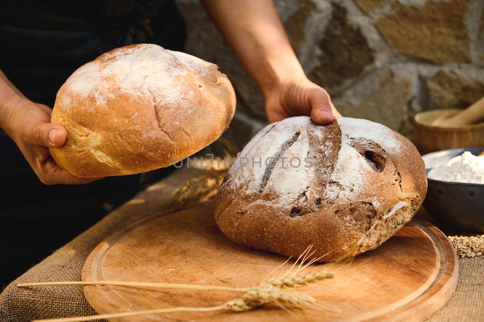 Details: baker hands hold various loaves of homemade whole grain and multigrain sourdough wheaten rye bread, over wooden cutting board with wheat ears. Healthy food, pastries enriched in dietary fiber