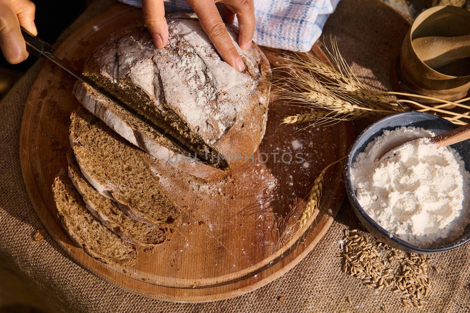 Overhead view of a baker slicing whole grain bread on a cutting board with a kitchen knife. A bowl of flour and spikelets of wheat, crumbs and wheat grains are scattered on the burlap tablecloth