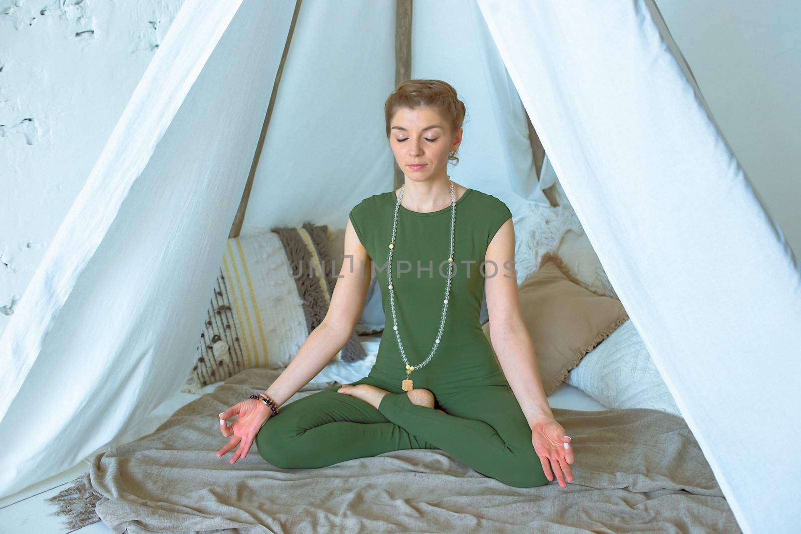 Beautiful and slender girl in green sportswear, meditates or practices yoga at home under a canopy of fabric