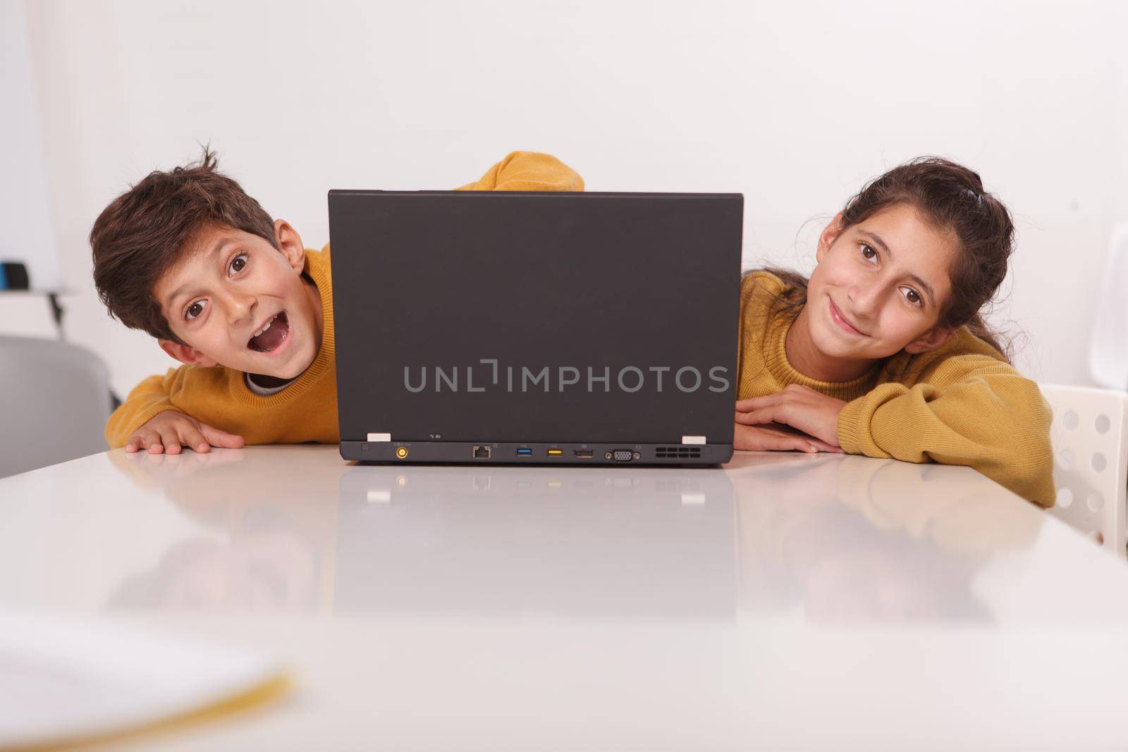 Cheerful brother and sister smiling to the camera, peeking from their laptop, copy space