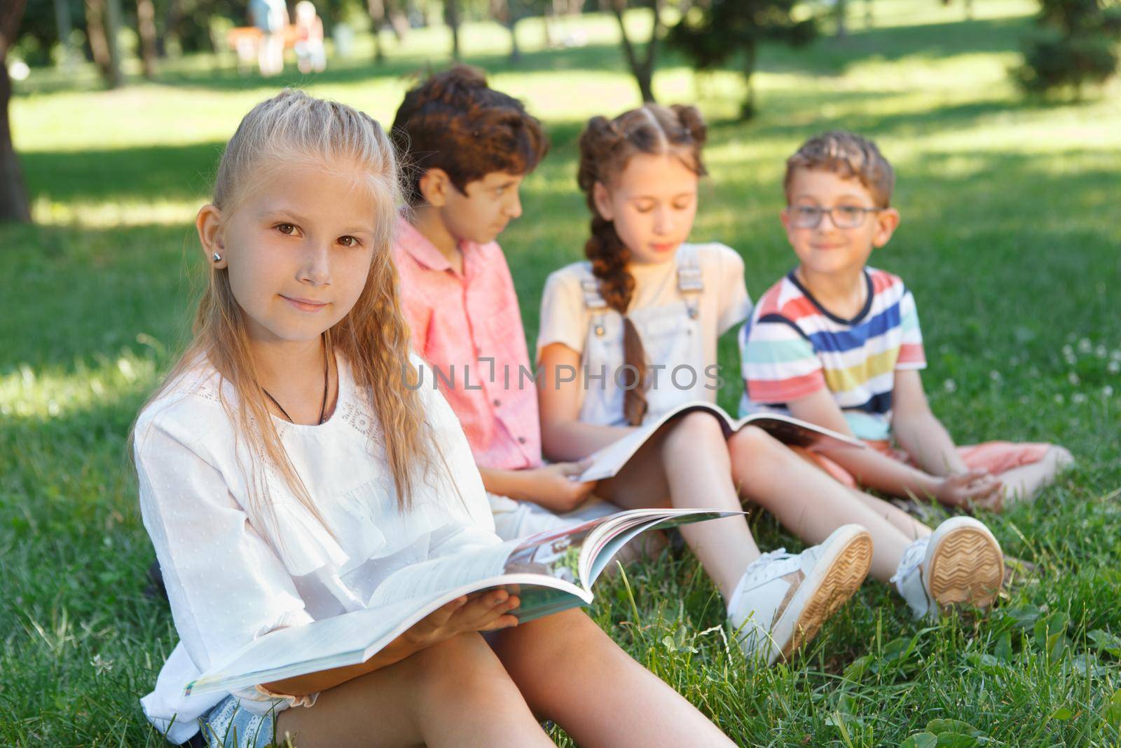 Lovely little girl reading a book at the park, her friends resting on the grass on background