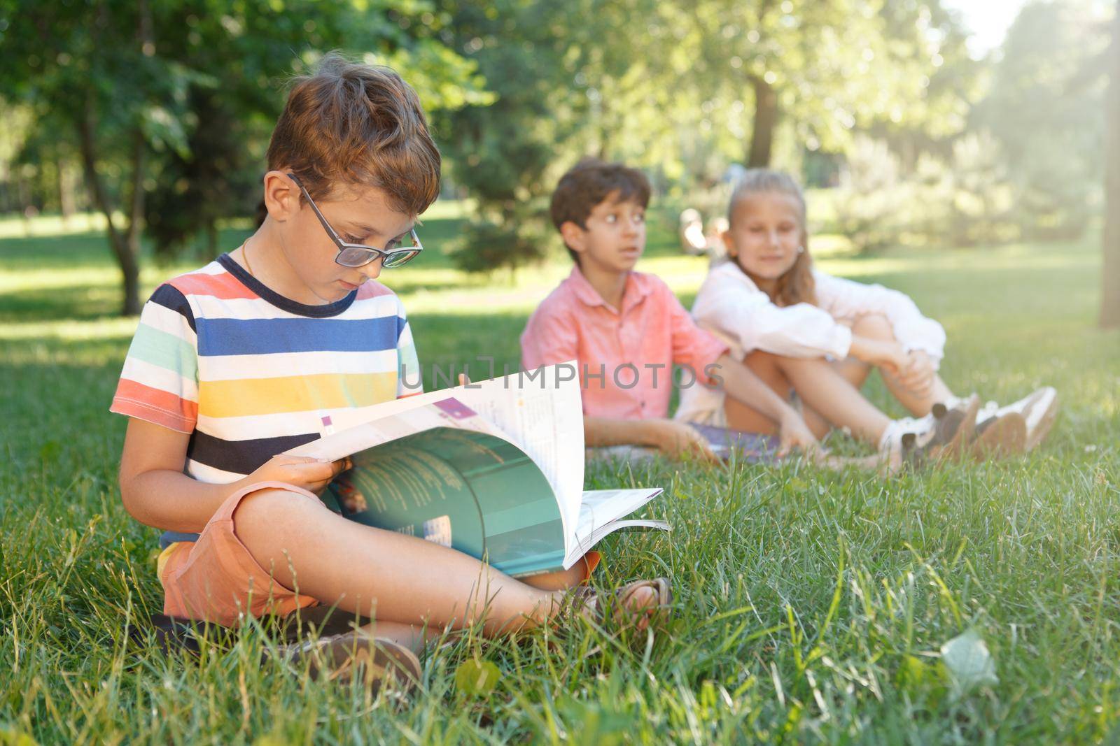Adorable little boy reading a book, sitting on the grass with his friends at the park