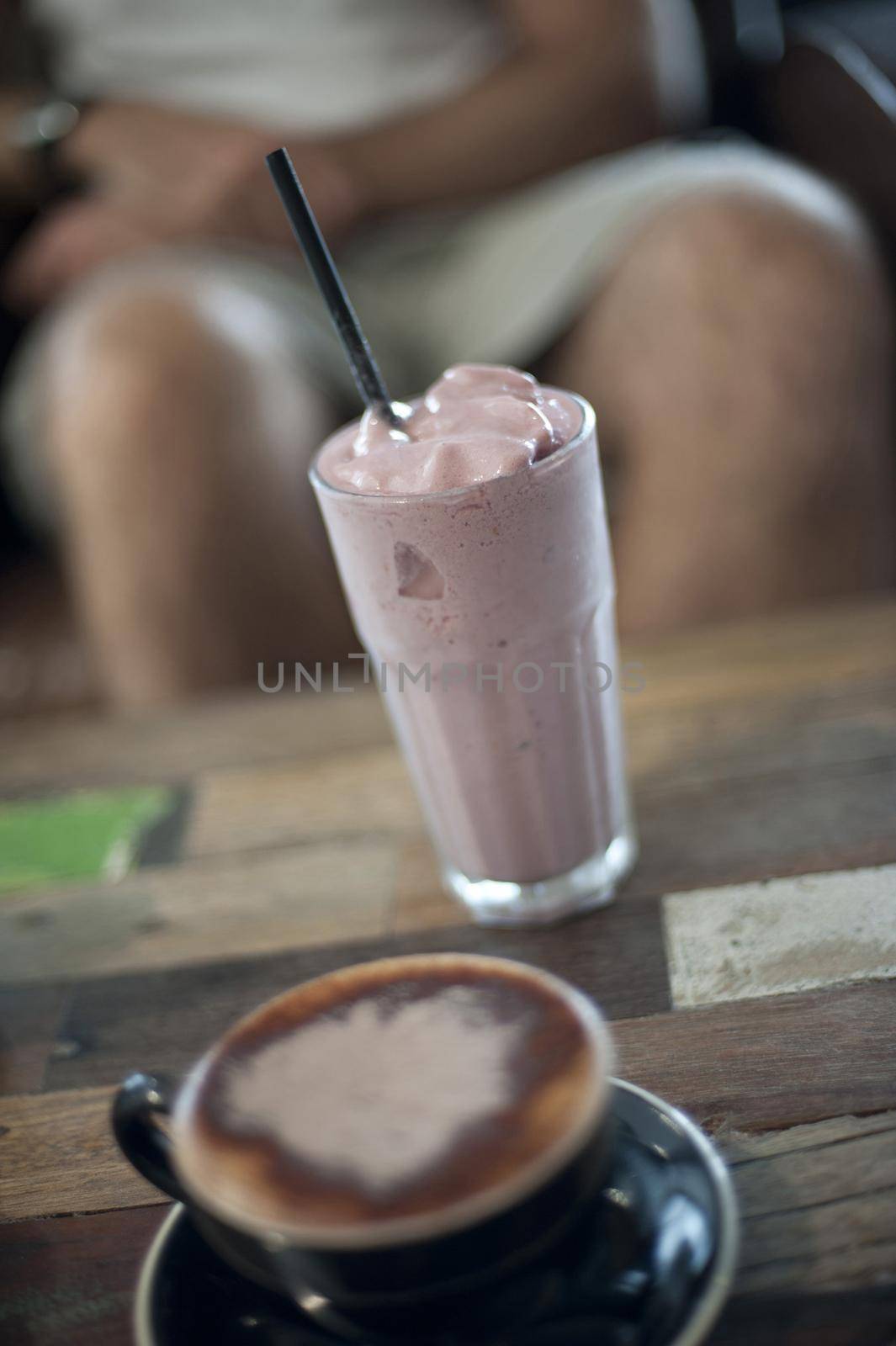 Fruit milkshake standing alongside a cup of cappuccino on a restaurant table, legs of a person seated at the table visible in the background