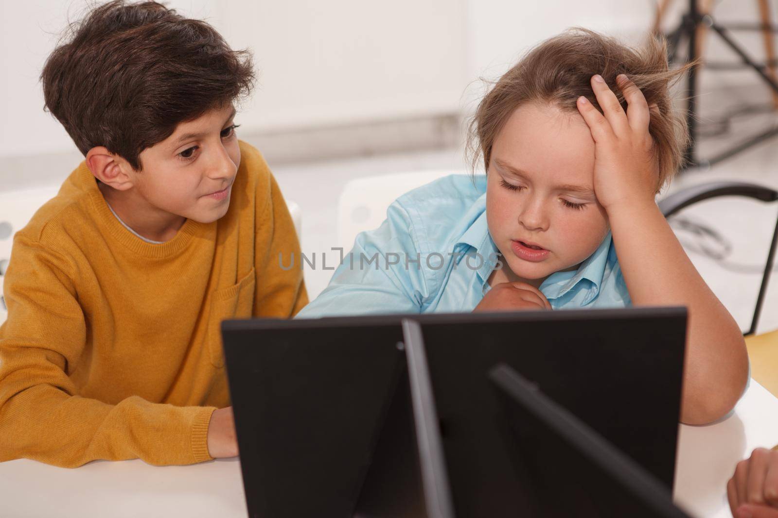 Adorable schoolboy looking tired, working on a school project with his friend, using laptop