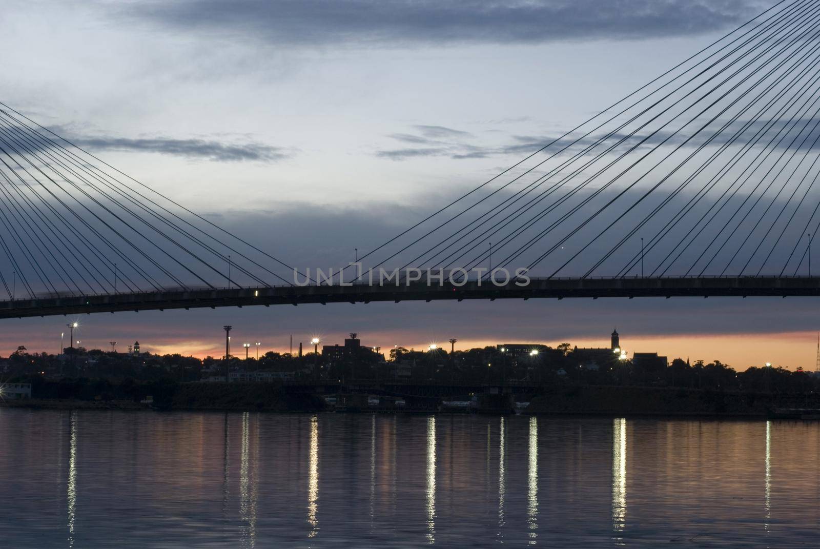 The middle of suspension bridge deck over water in twilight with the lights reflecting in calm water surface