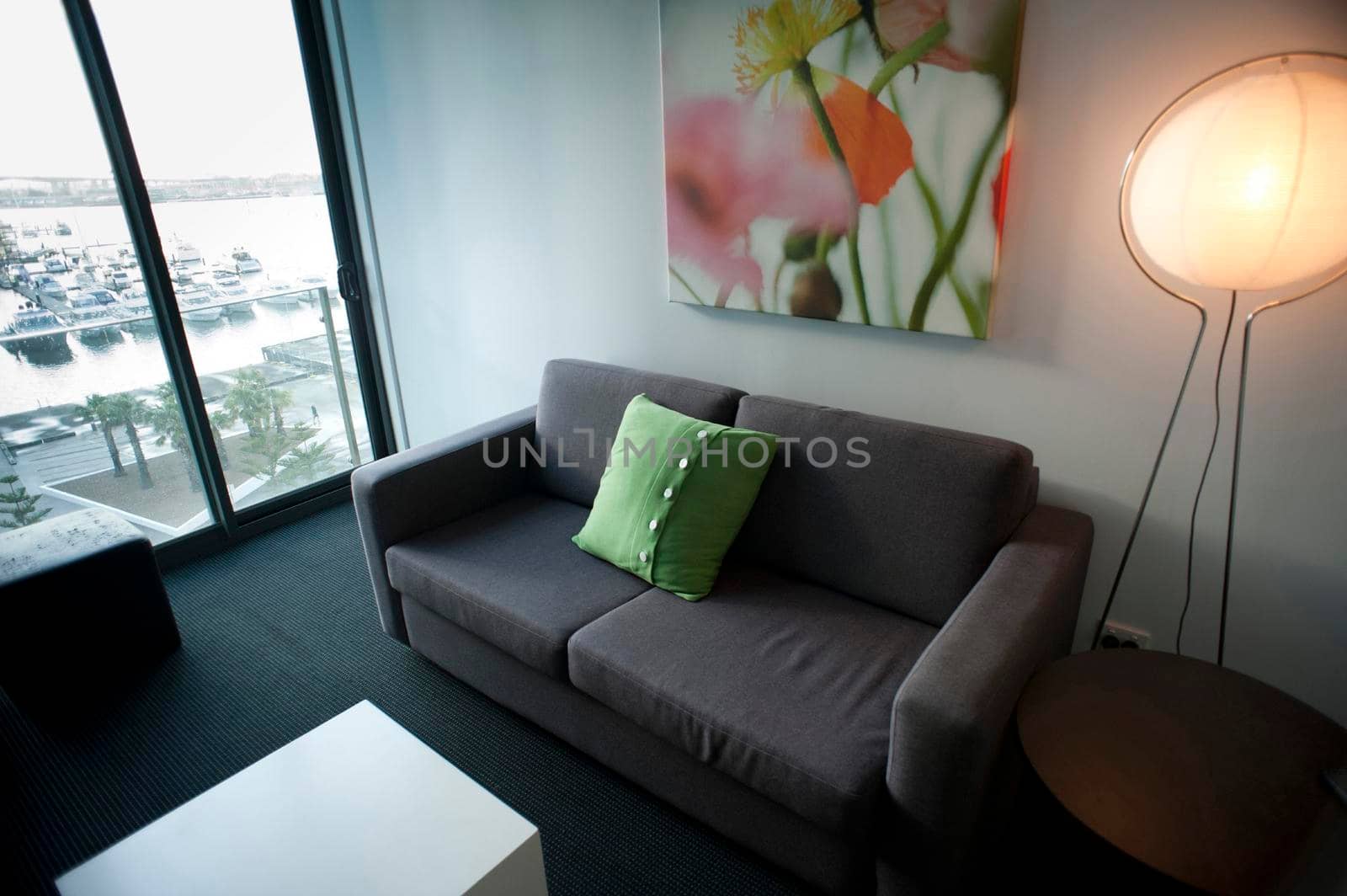 Interior decor of a compact city apartment with a modern lounge with an illuminated standard lamp alongside a grey upholstered couch with modern artwork and a view window