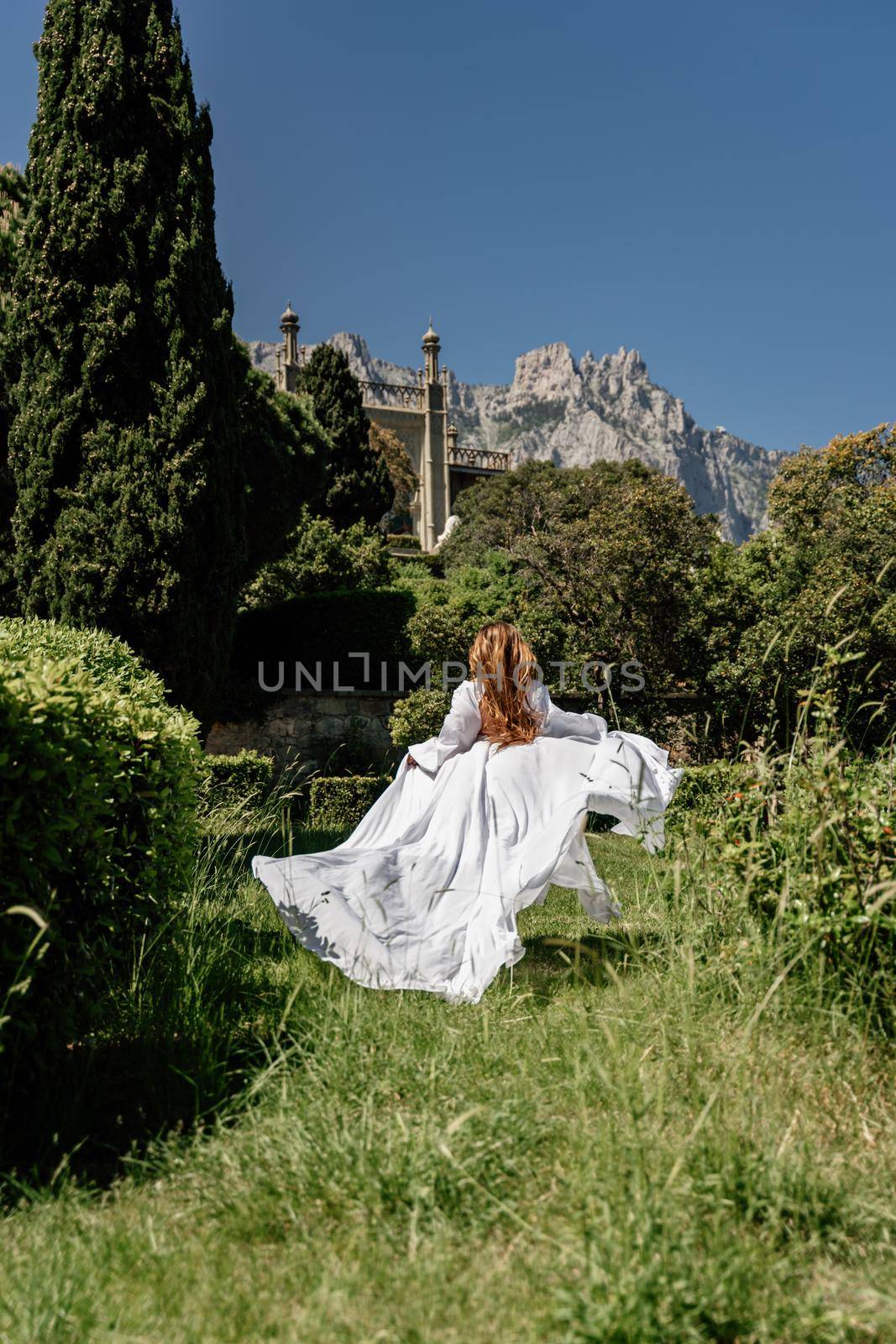 a beautiful woman with long brown hair and long white dress stay along a path along beautiful bushes in the park rear view by Matiunina