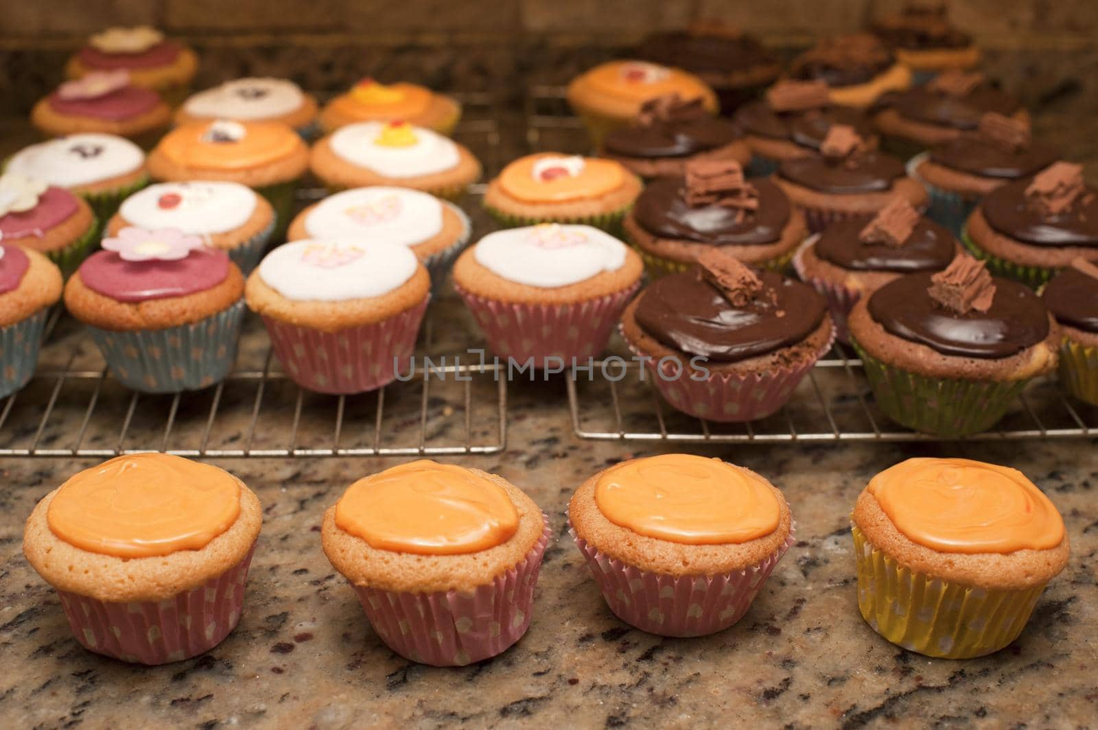 Making a batch of fairy cakes or cupcakes in the kitchen decorating them with colorful icing and flowers as they stand on wire trays on the counter cooling after baking