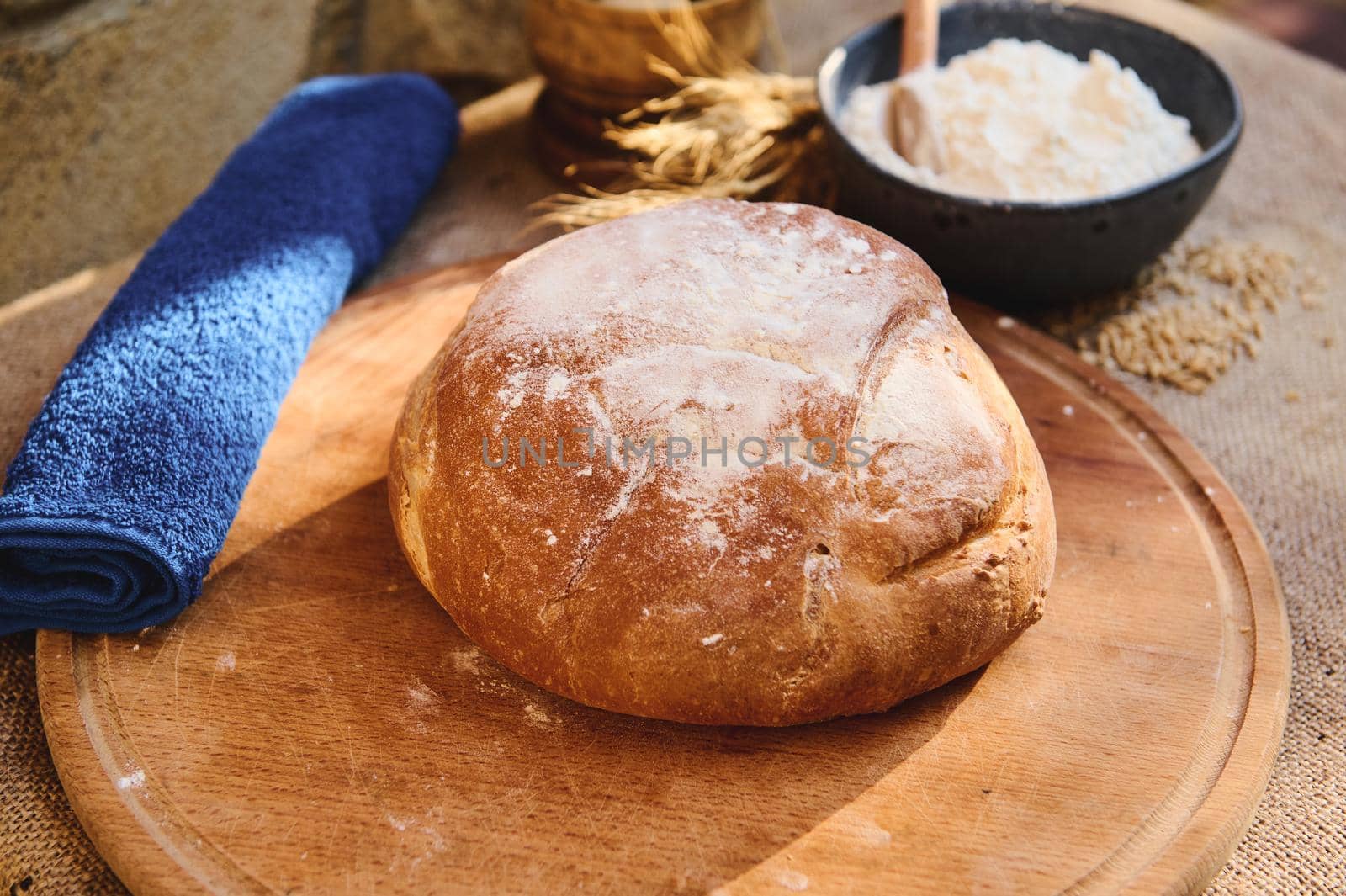 Still life with a loaf of homemade whole grain sourdough bread, freshly baked according to family traditions, on a wooden board, a blue terry towel and a bowl with wheat flour. Artisanal bakery store