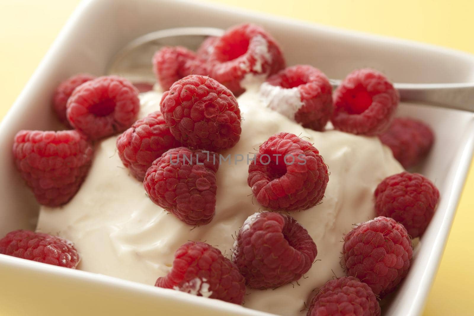 Plain creamy yogurt topped with fresh raspberries in a box shaped bowl with spoon