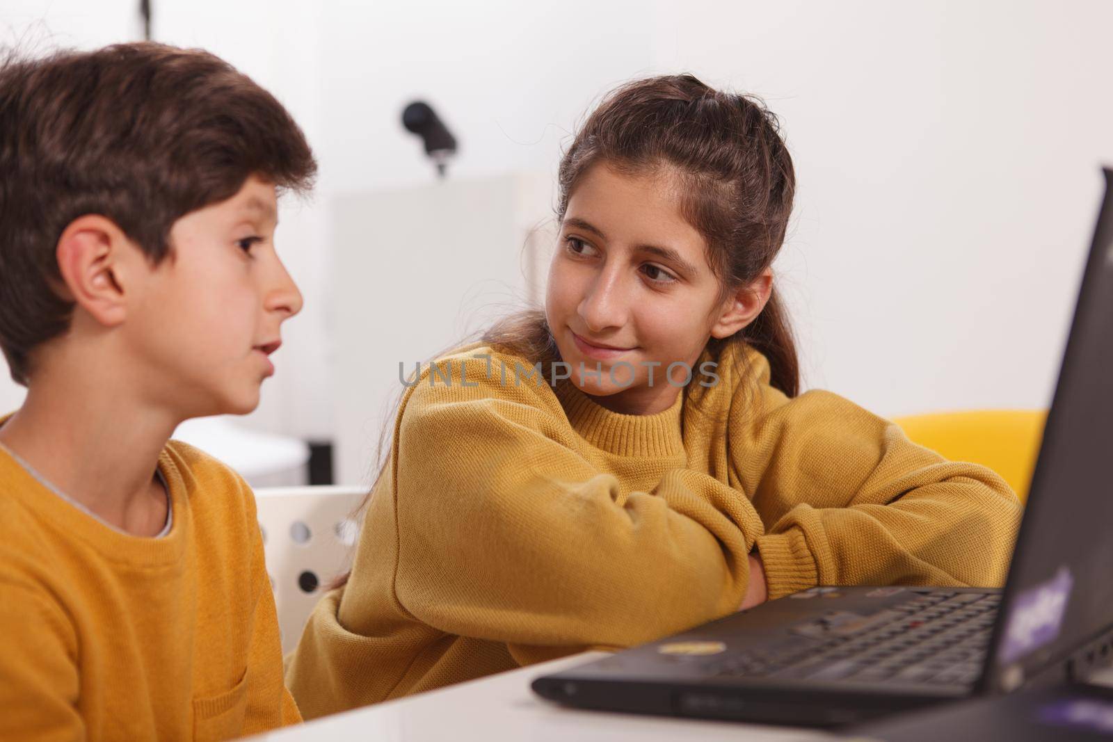 Lovely teen Arab girl talking to her little brother while doing homework together on a laptop