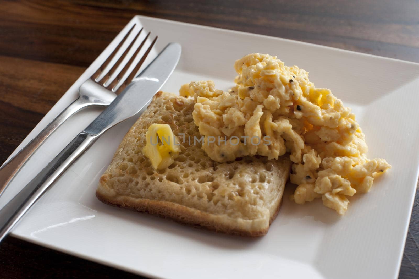 Close up view of scrambled eggs with a crumpet besides fork and spoon on a square white plate