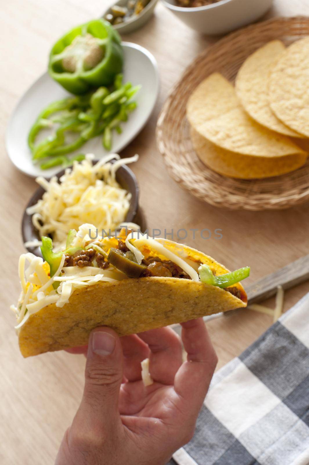 Selective focus of hand holding hard shelled taco beside bowl of shredded cheese and plate of cut green bell pepper