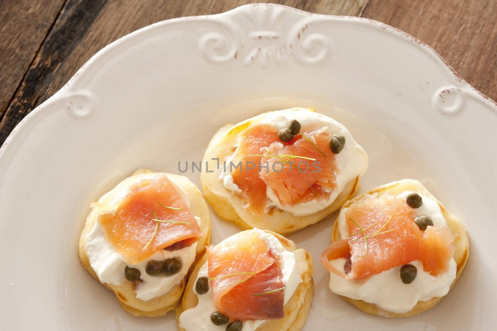 Capers and raw fish blini appetizers on white porcelain decorative plate over wooden table