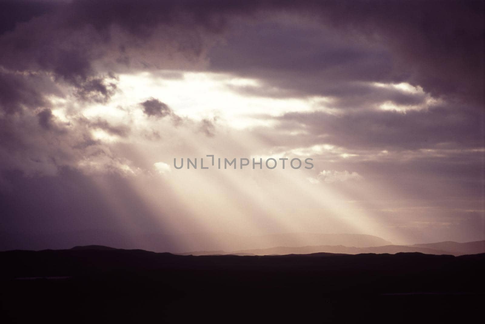 Sunbeams shining through a hole in the dense cloud cover in a stormy grey sky throwing diagonal rays onto the silhouetted hills below