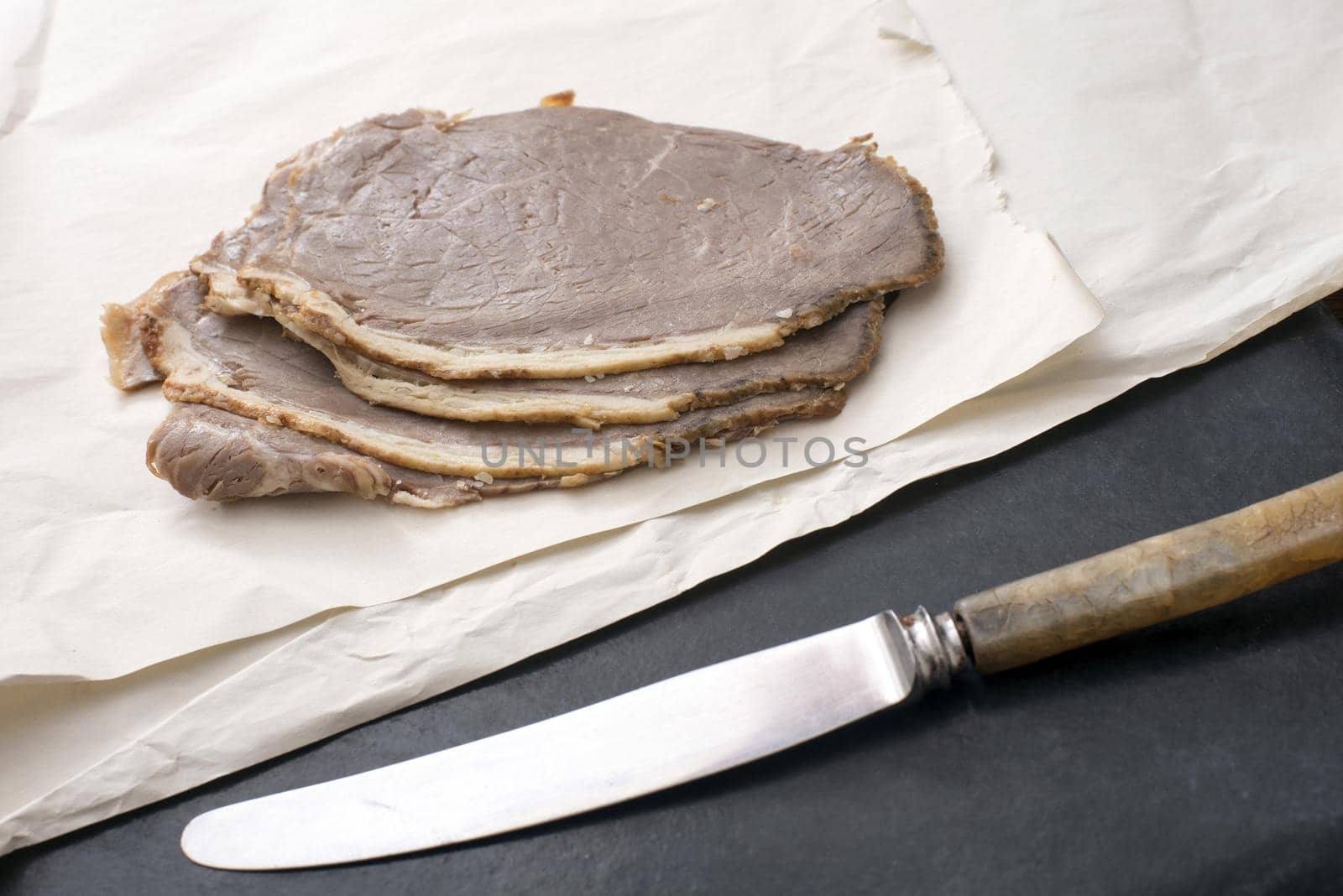 Pile of cold sliced roast beef for sandwiches on crumpled white paper with a kitchen knife in the foreground