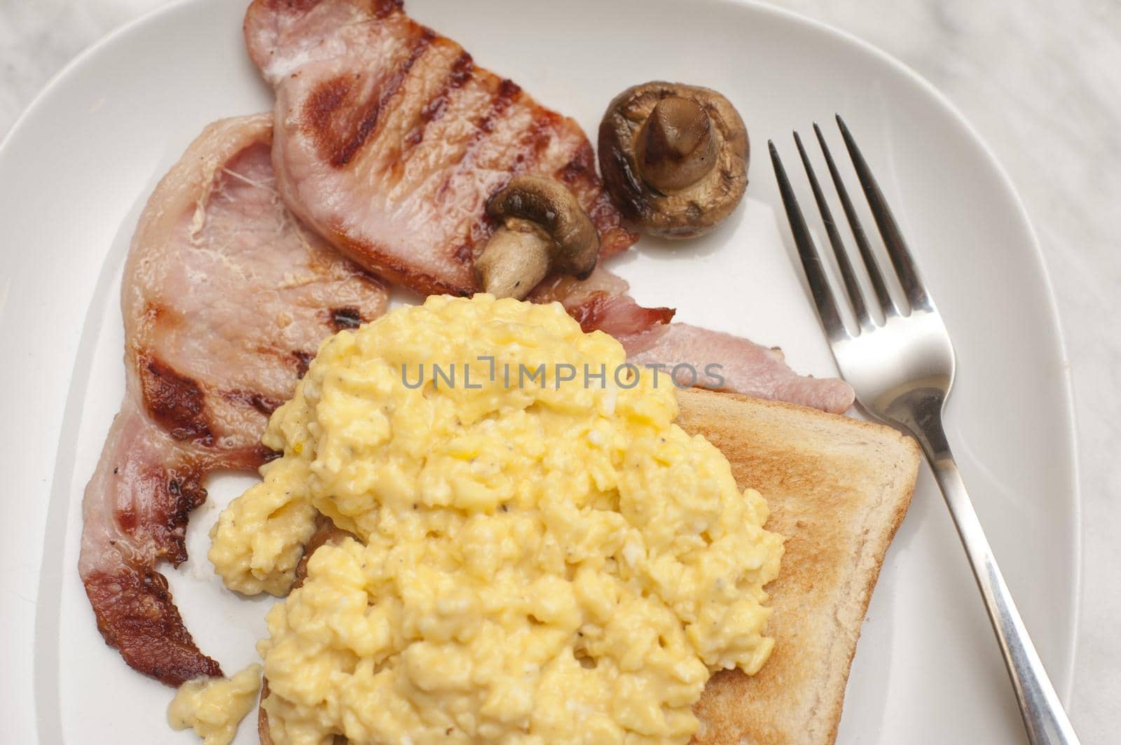 Scrambled egg on toast served with lean grilled bacon rashers and mushroom , close up view from above