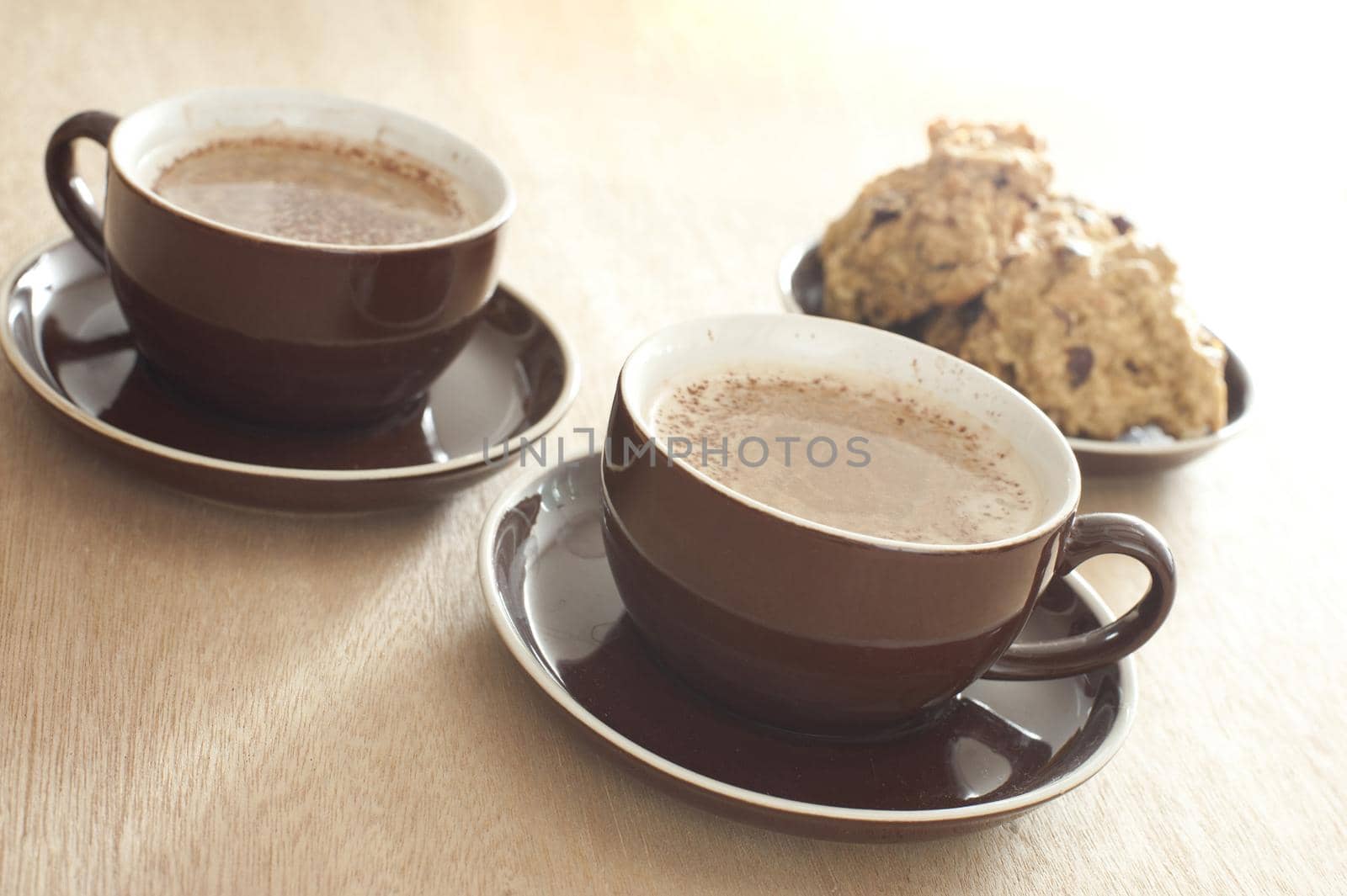 Coffee time with two generic brown cups and saucers of frothy fresh coffee served with a plate of sliced cake, focus to the cup in the foreground