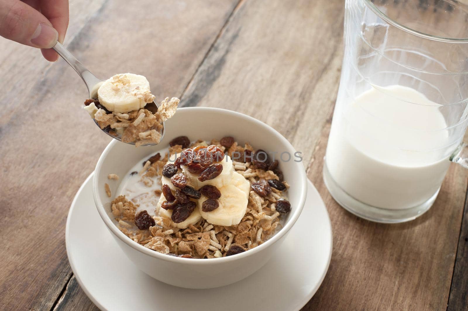 First person perspective view of spoon in hand reaching up to eat muesli granola cereal with slices of banana and raisins on top beside half empty glass pitcher of milk over table
