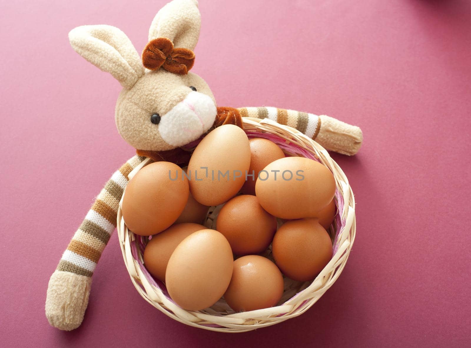 Cute toy Easter bunny with striped arms around a wicker basket filled with fresh brown eggs over a red background