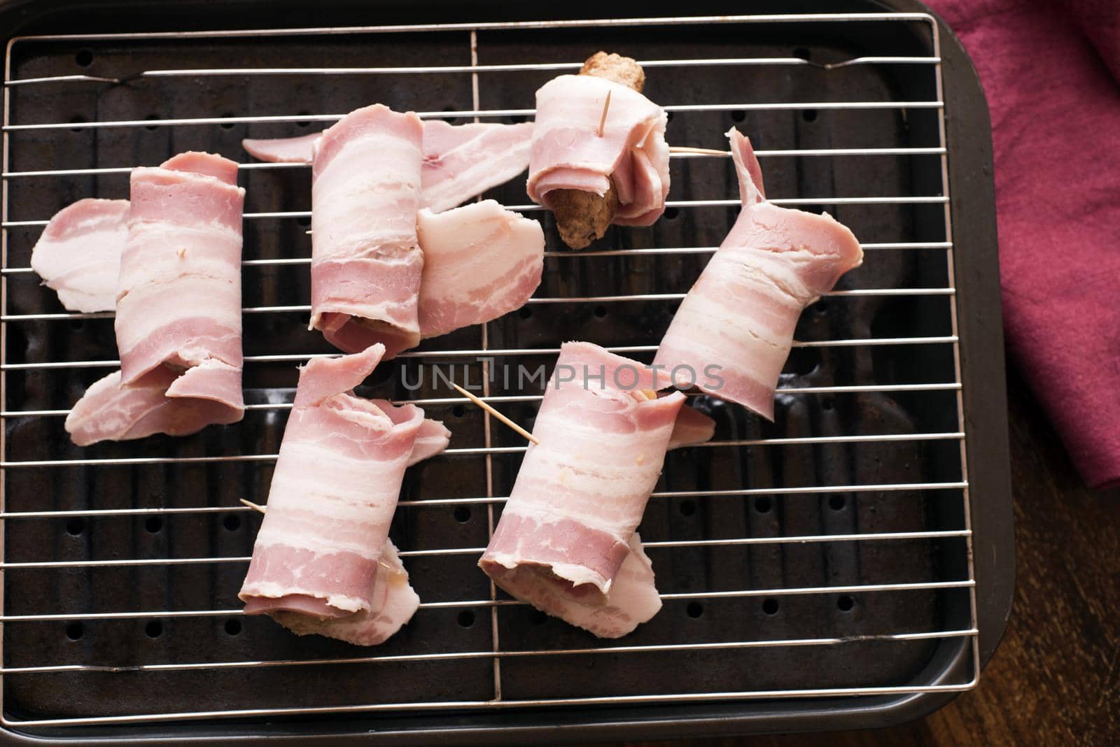 Making a batch of pigs in blankets or bacon rolls around pork sausages laid on a grill pan ready to place in the oven viewed from above