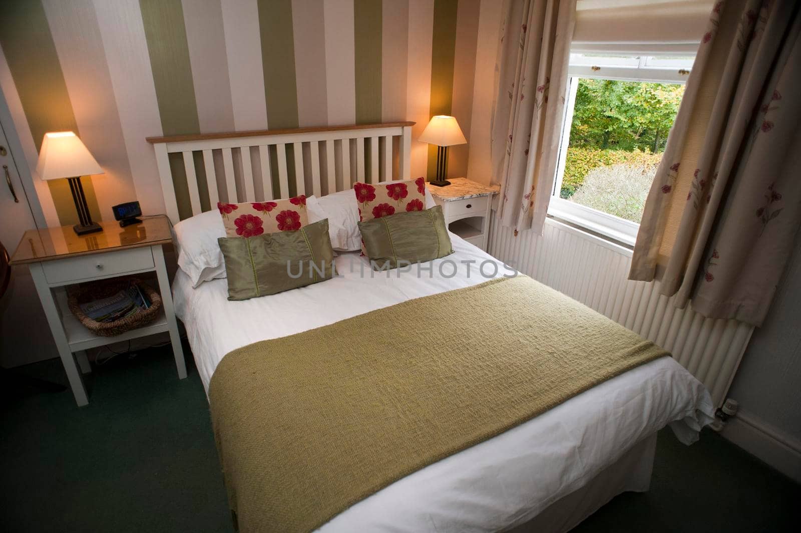 Homely bedroom interior with a double bed and lighted lamps on bedside tables alongside a window with a garden view and striped wallpaper
