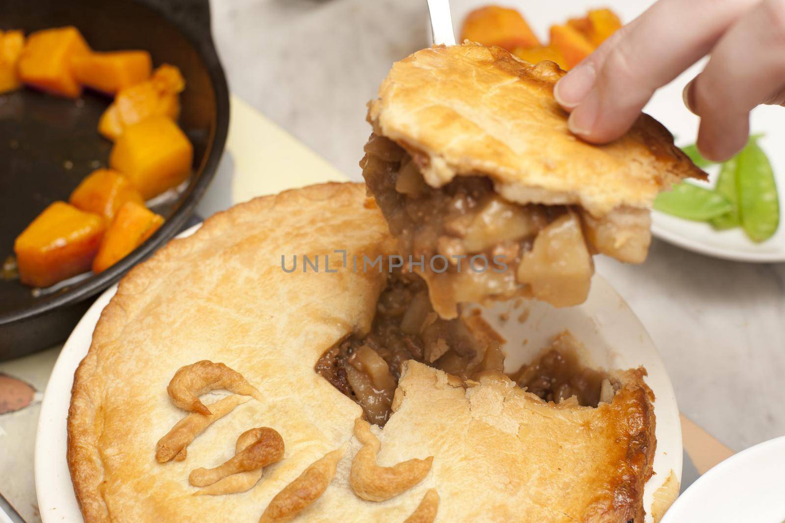 Person serving a tasty homemade meat and potato pie with a golden pastry crust spooning a portion onto a plate with vegetables