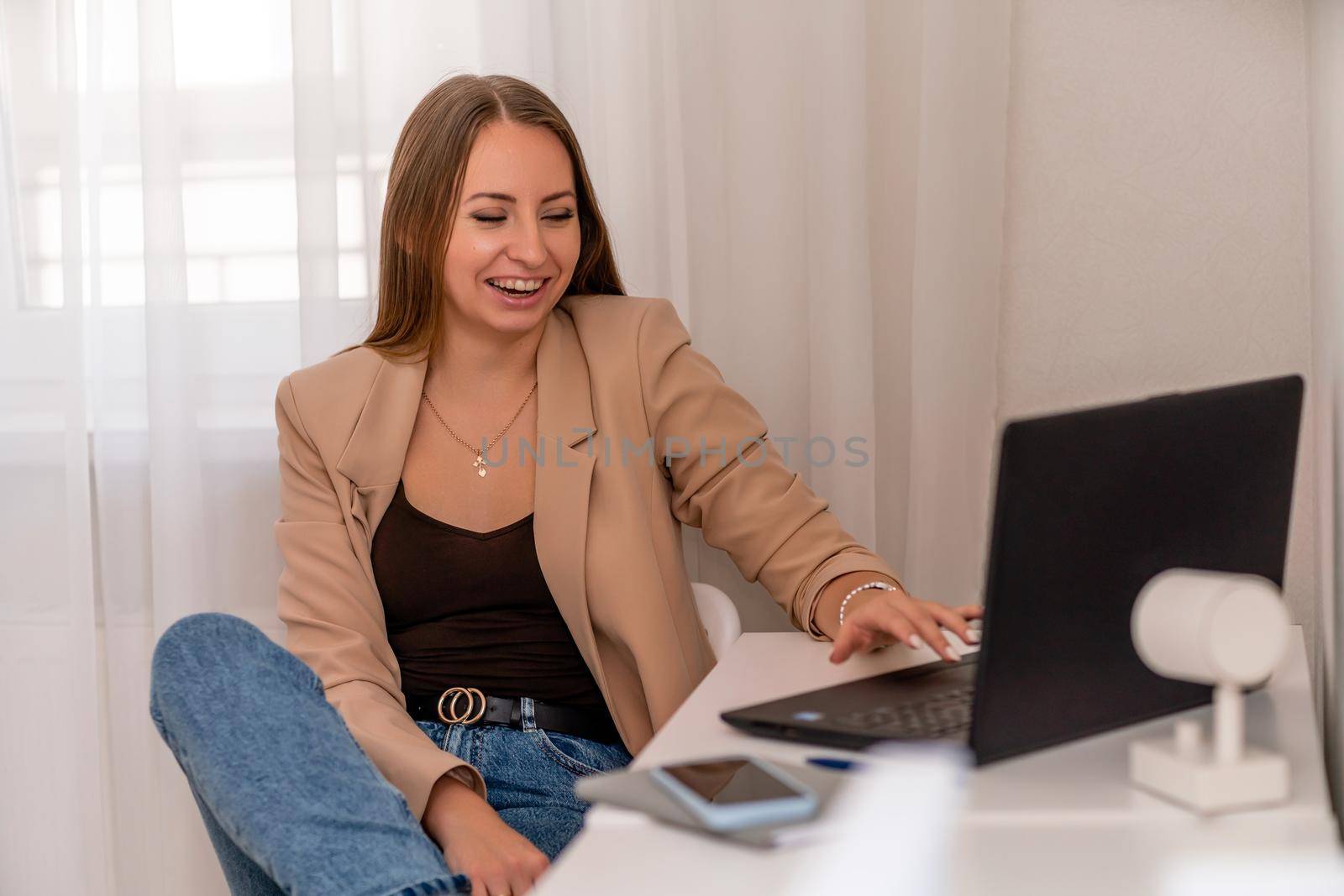 European professional woman sitting with laptop at home office desk, positive woman studying while working on PC. She is wearing a beige jacket and jeans