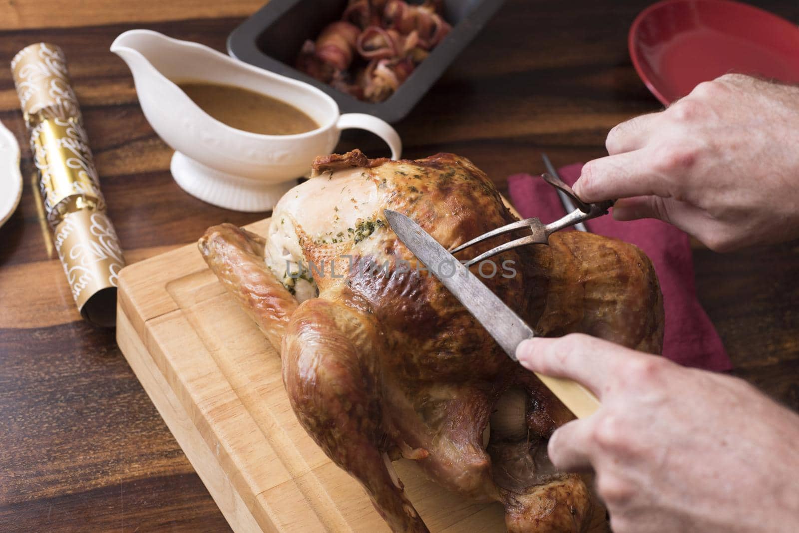 Man carving a Christmas turkey with vintage bone handled utensils on a wooden cutting board on a decorated table with gravy in a sauce boat