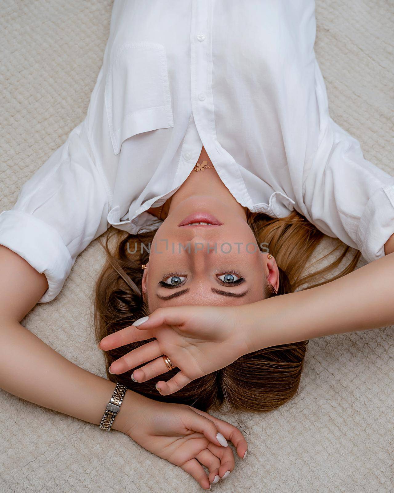 The woman lies on the bed on her back, top view. She looks straight ahead, wearing a white shirt by Matiunina