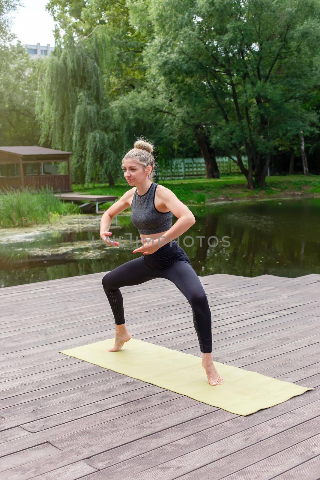 A slender woman in a black top and leggings does flow yoga in summer, standing on a wooden platform by a pond in the park. Copy space. Vertical