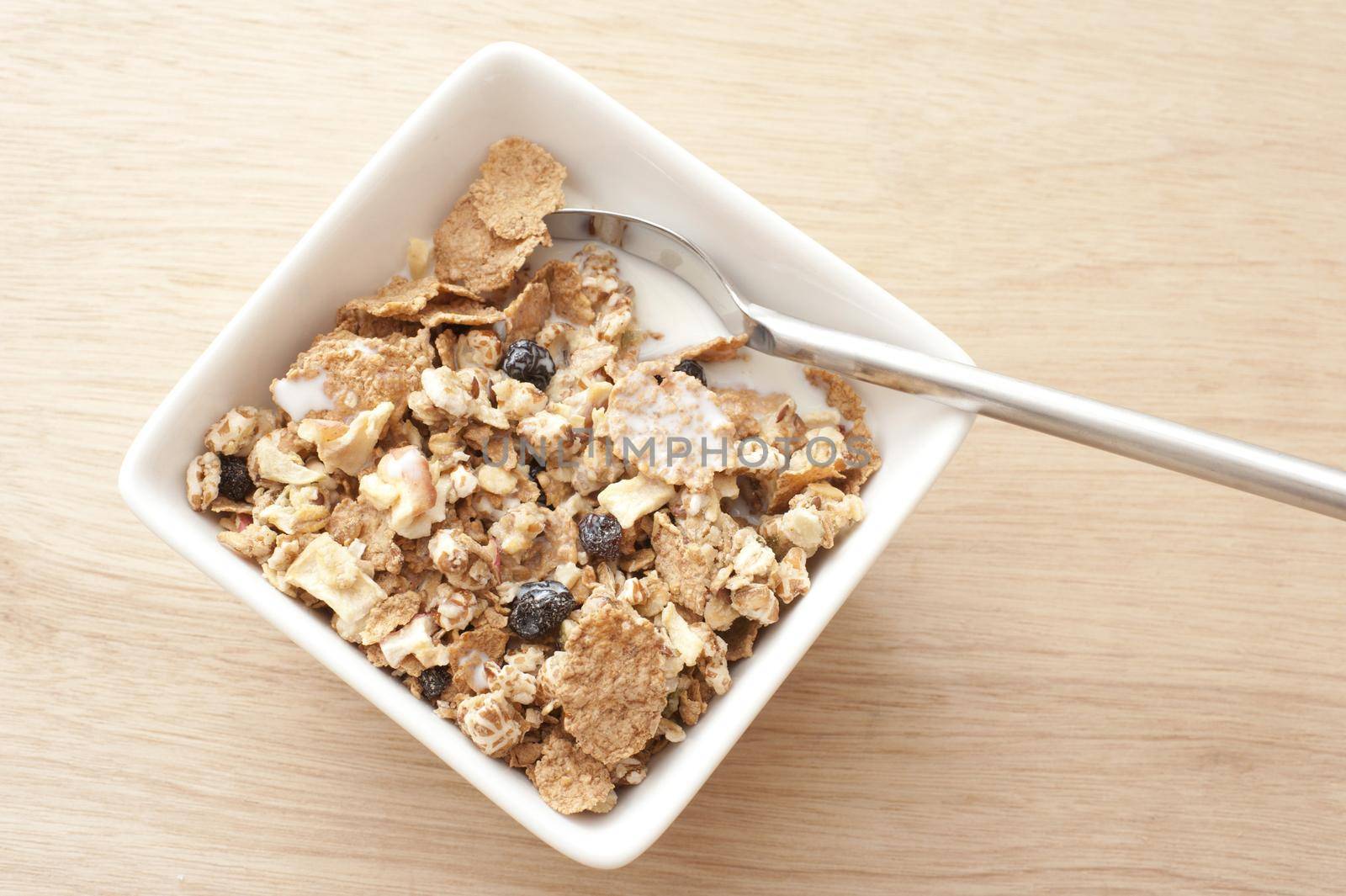 First person perspective view of spoon in square bowl of muesli granola cereal with slices of dried fruit on top with milk over wooden table