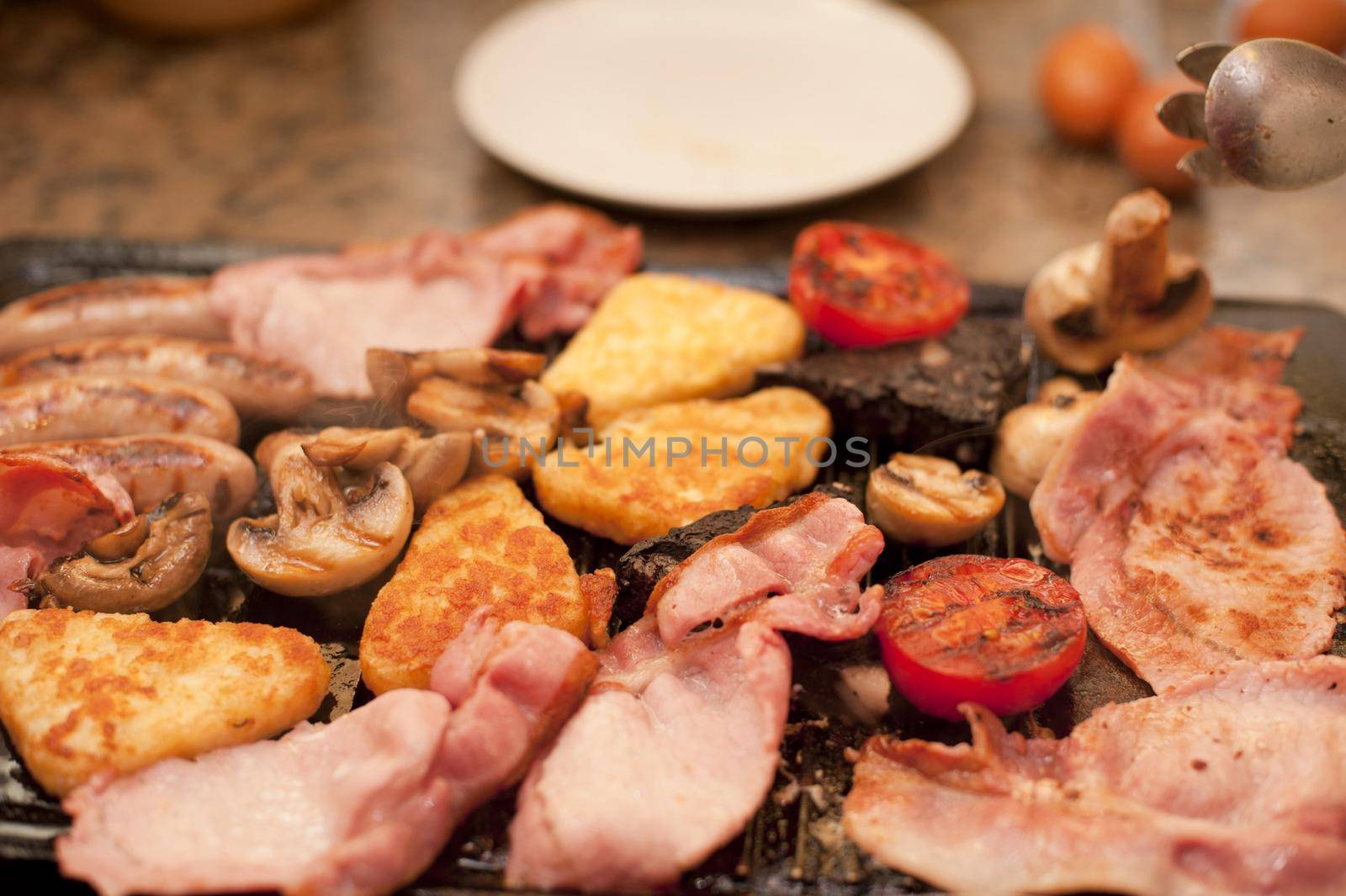 Wholesome English breakfast cooking on the griddle with bacon, hash browns, sausages, tomatoes and mushrooms