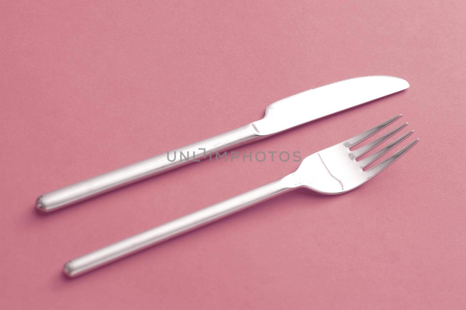 Simple design silver knife and fork lying side by side on a red background diagonally across the frame