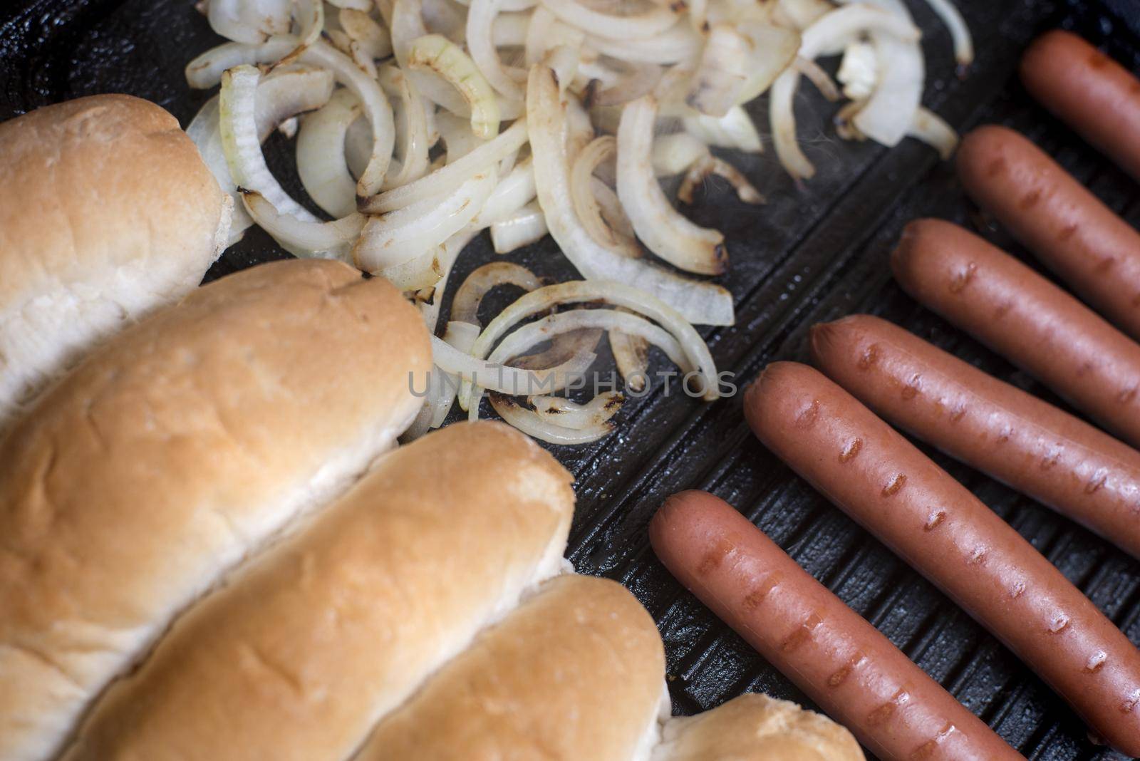 Cooking hot dog ingredients on a griddle with a row of smoked Wiener or Frankfurter sausages, diced onion and fresh white bread rolls