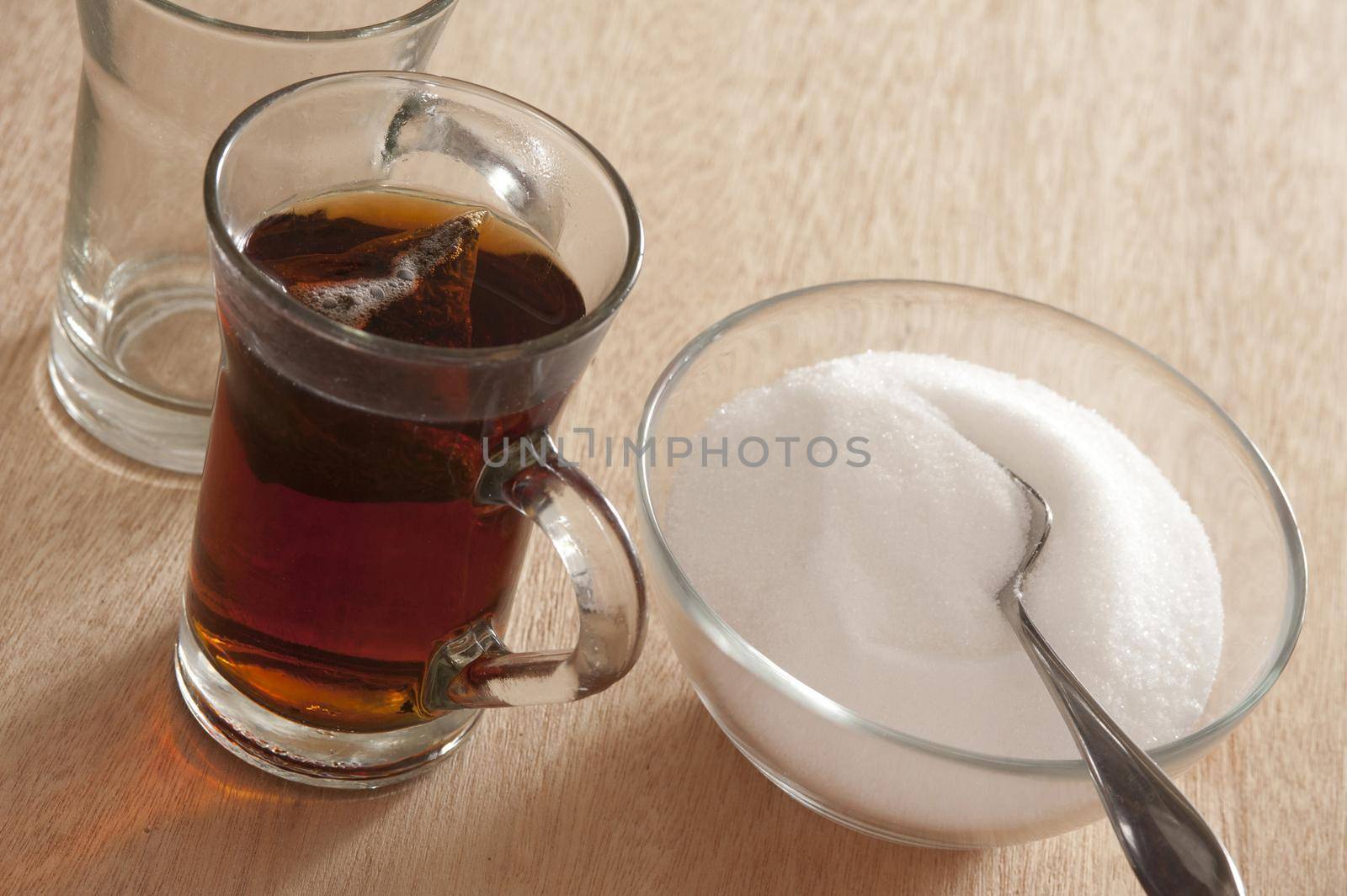 Sugary sweet hot black tea in a glass mug with a bowl full of white sugar and a teaspoon alongside on a wooden table, high angle view