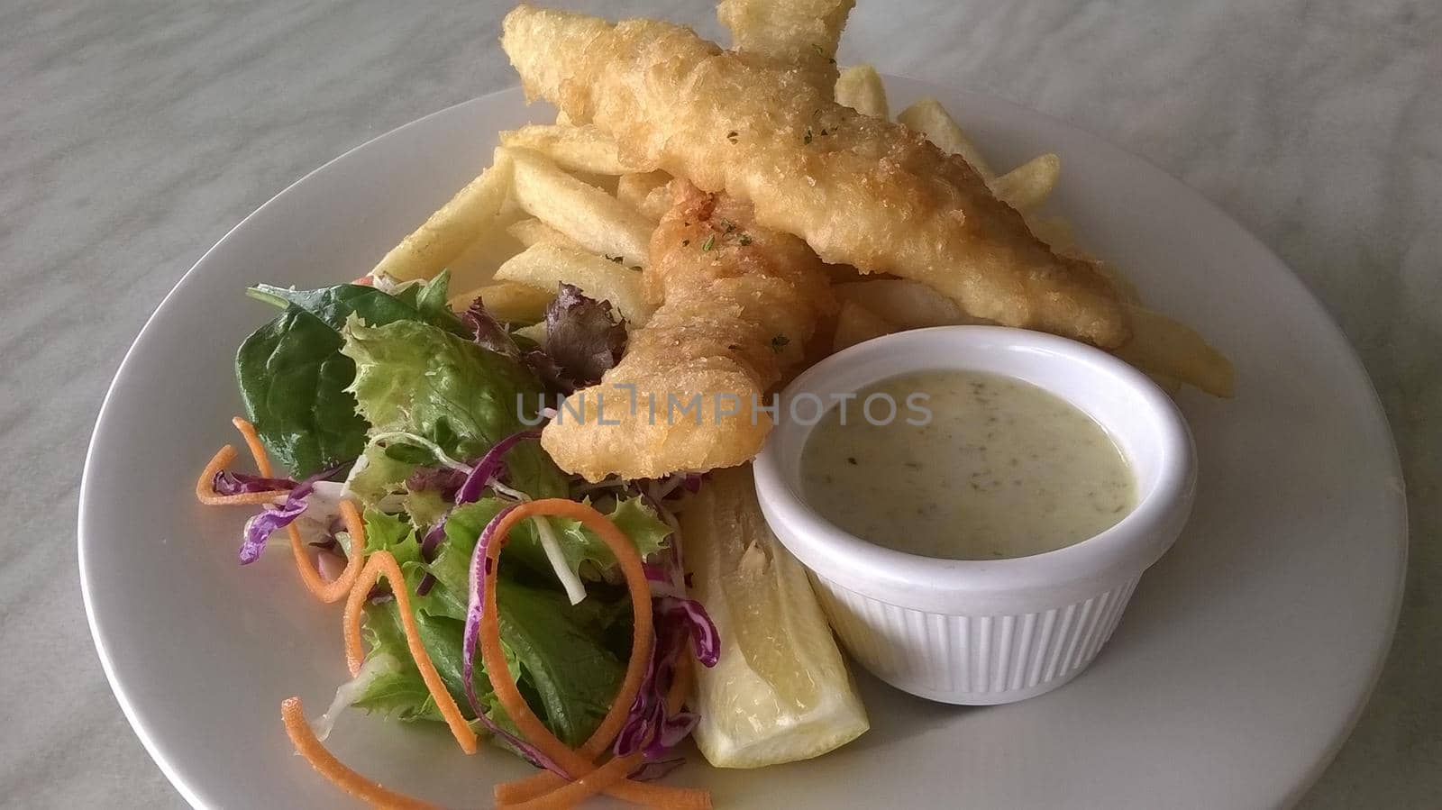 Battered fish and fried potato chips served with a savory sauce and a fresh salad on a white plate, close up high angle view