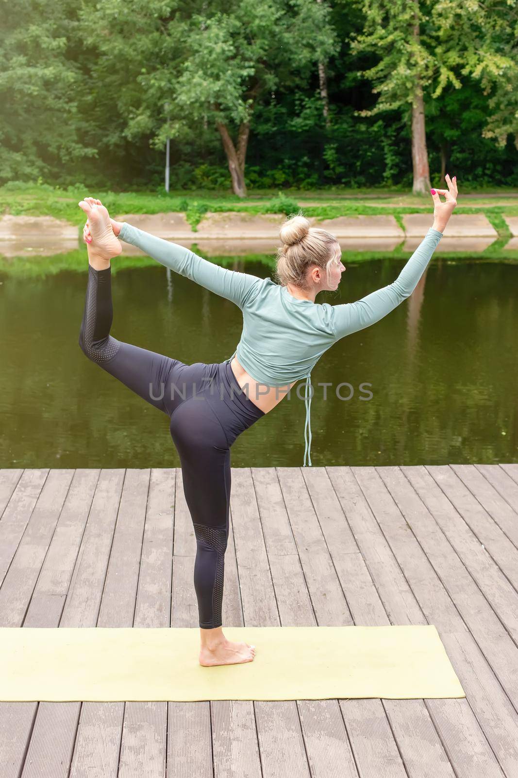 A slender woman in a green top and leggings does flow yoga in summer, standing on a wooden platform by a pond in the park. Vertical